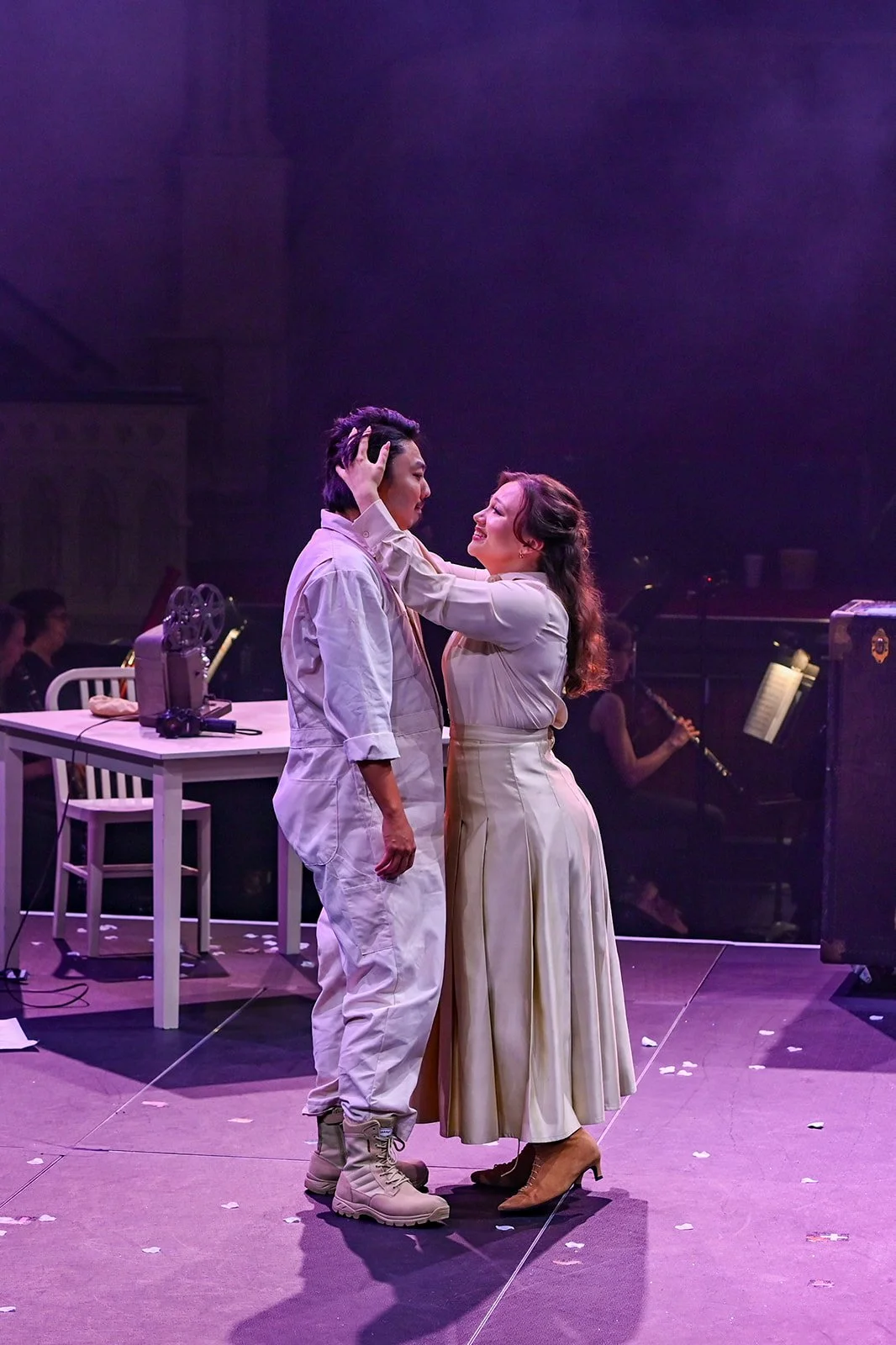 Two actors on stage, a man and woman, share an intimate moment, looking into each other's eyes. The woman touches the man's face. In the background, musicians and a piano can be seen, with stage lighting creating a dramatic atmosphere.