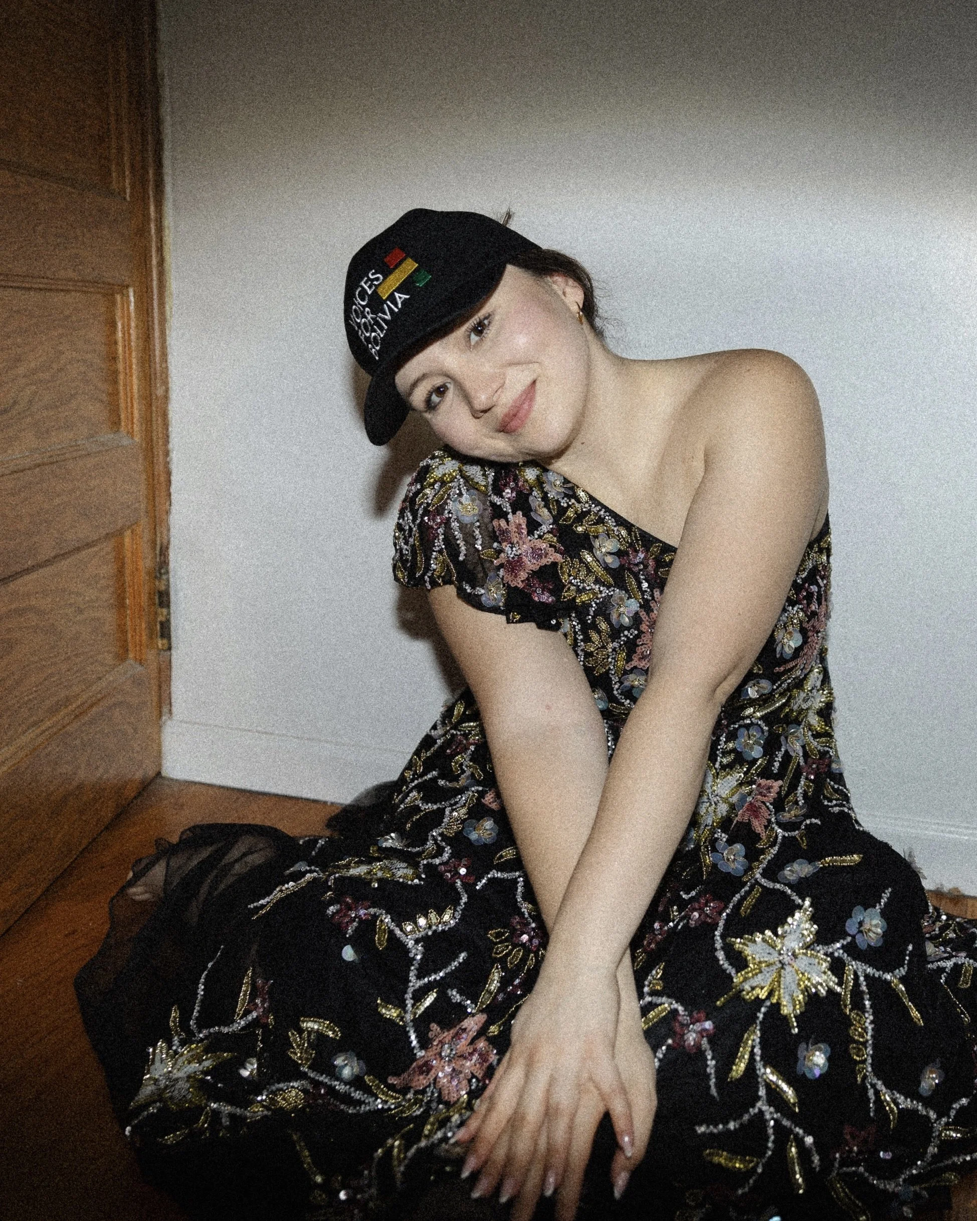 A woman with fair skin and makeup, wearing a black hat with colorful embroidery and a floral dress with intricate beading, sitting on the floor, showing her smiling face and resting her head on her arm. The woman is near a wooden door and a white wal