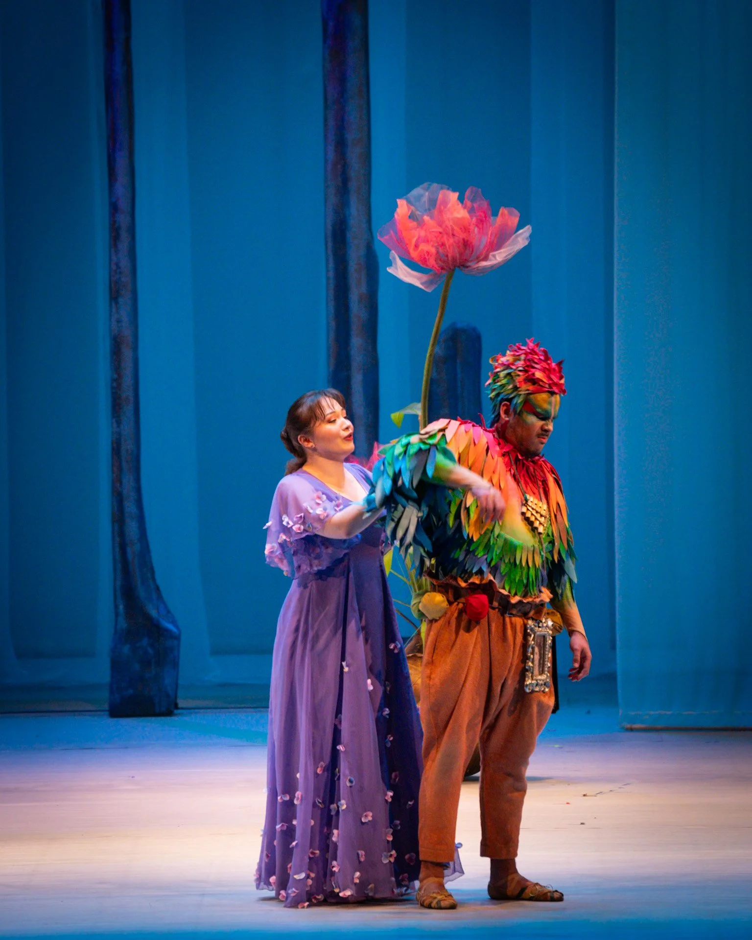 Two performers on stage, one woman in a purple dress holding the hand of a man in colorful costume with a large pink flower headpiece, against a blue background.