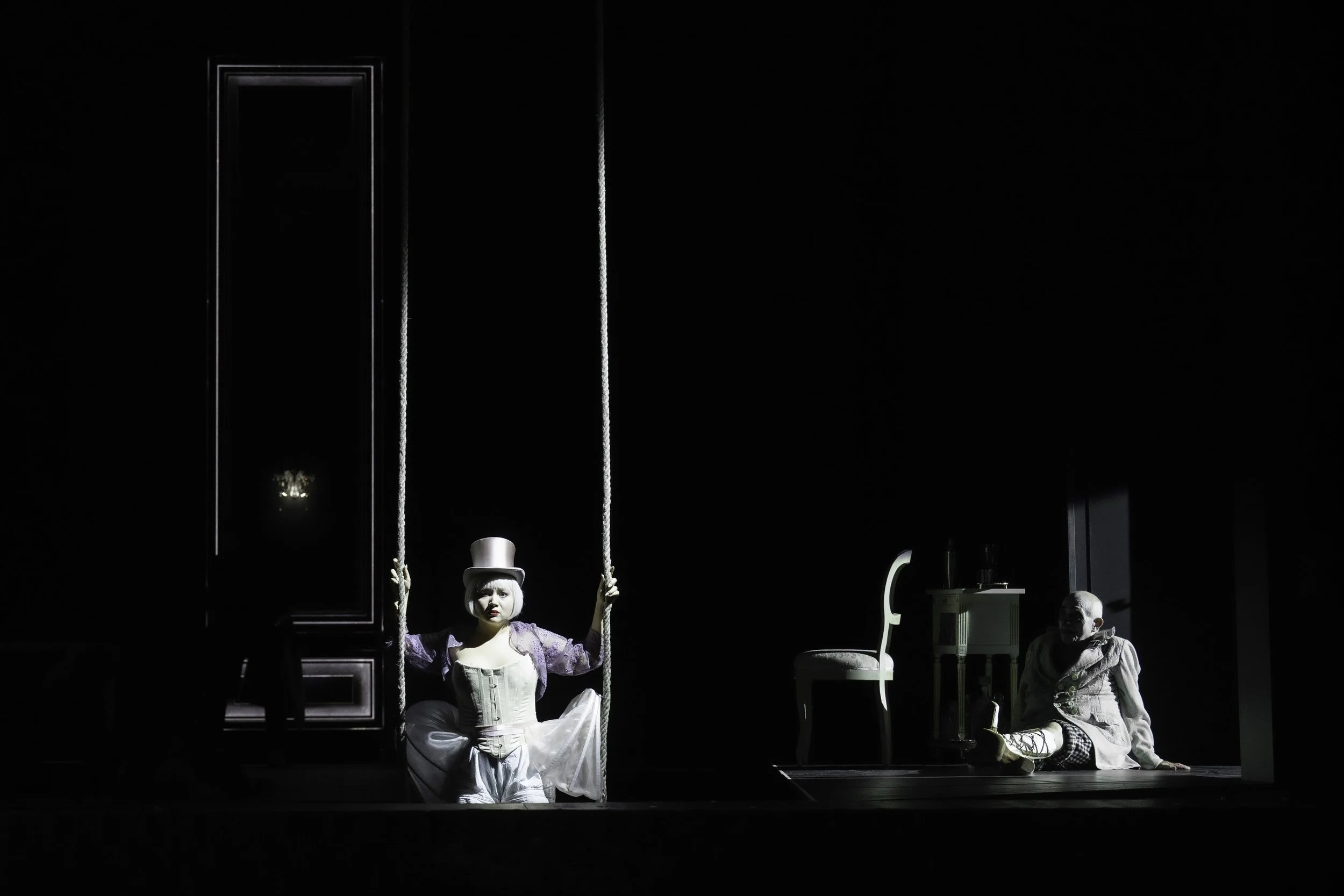 A theatrical performance featuring two characters: a woman sitting on a swing in an elaborate costume and a man sitting on the floor nearby in casual attire, with a dark background and minimal set pieces.