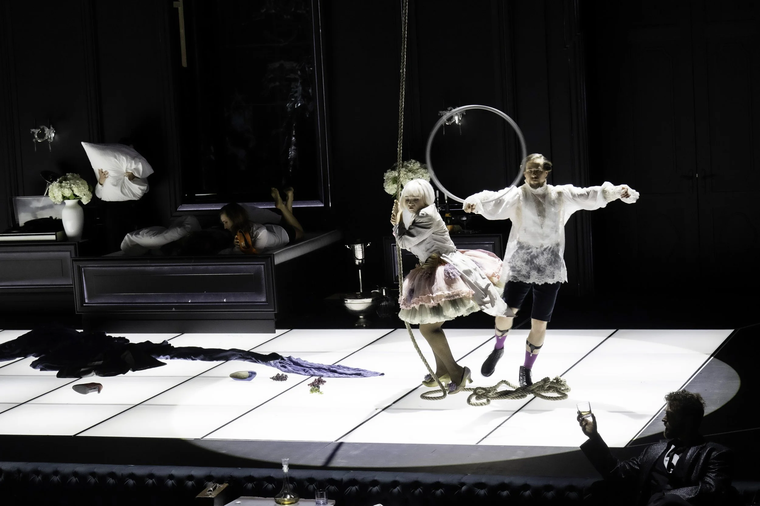 A theatrical stage scene with performers in elaborate costumes. One performer is dressed as a doll with a pink tutu and blonde wig, holding a rope, and another is dressed in lace with a hoop, sparring with the doll. In the background, a person appear