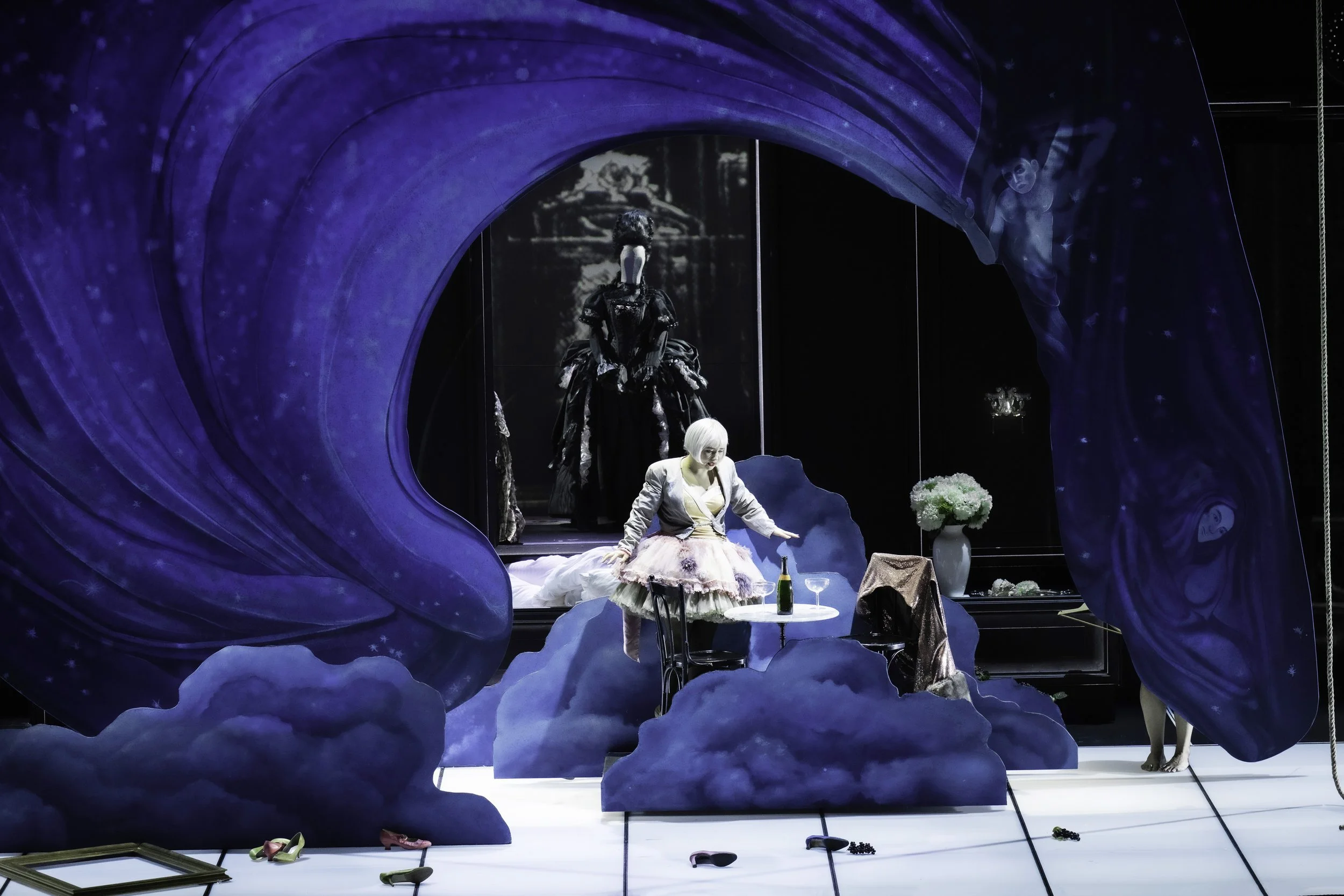 A theatrical scene with a woman in a pastel-colored dress sitting at a small table with a bottle, glasses, and flowers, surrounded by dark blue clouds and two women in elaborate black and white costumes in the background. The stage is decorated with 