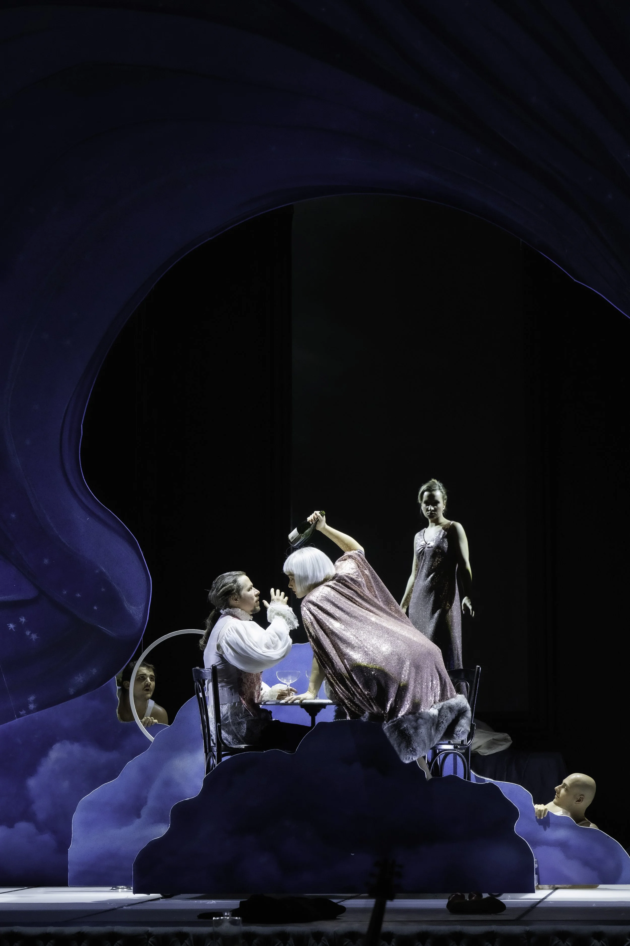 A theatrical stage scene with actors and mannequins surrounded by cloud-like props against a dark background, with a large blue structure on the left.