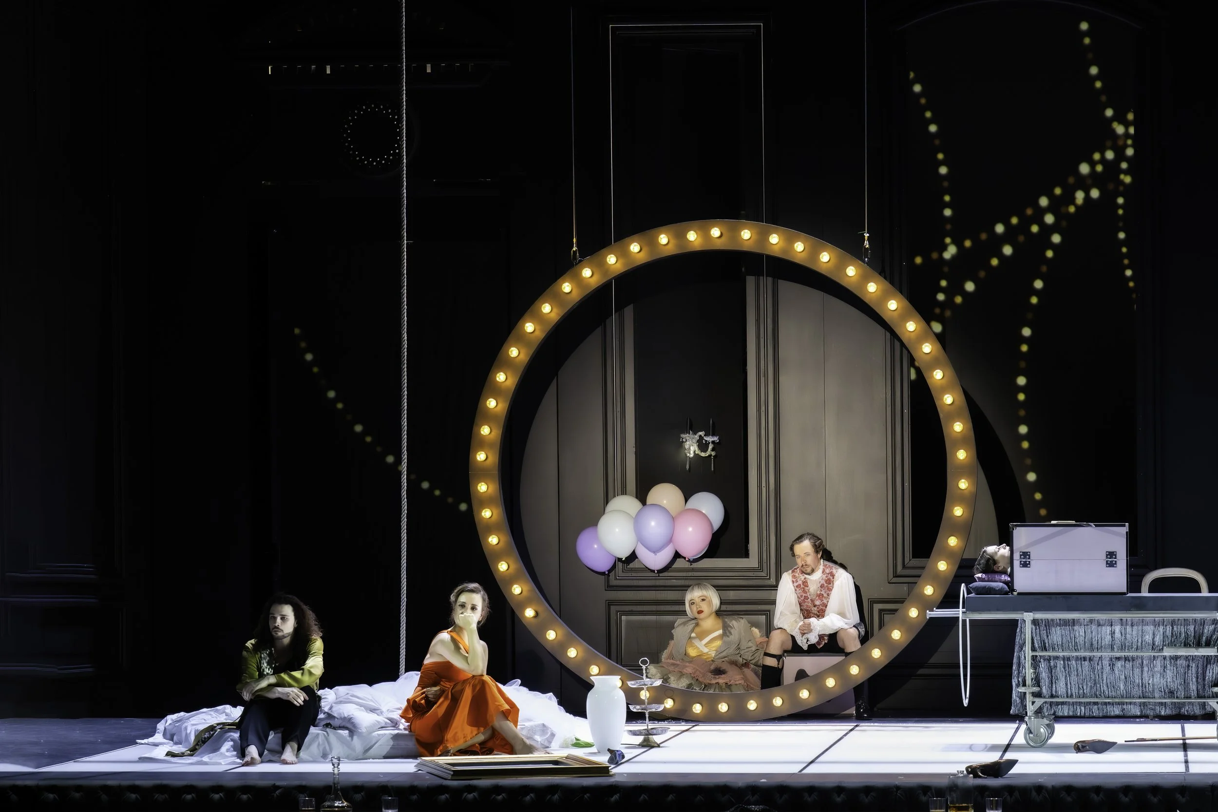The image shows a stage scene with five performers, three women and two men, seated or standing amidst props including balloons, a white vase, and books. There is a large illuminated circular frame and a backdrop with dotted light patterns.