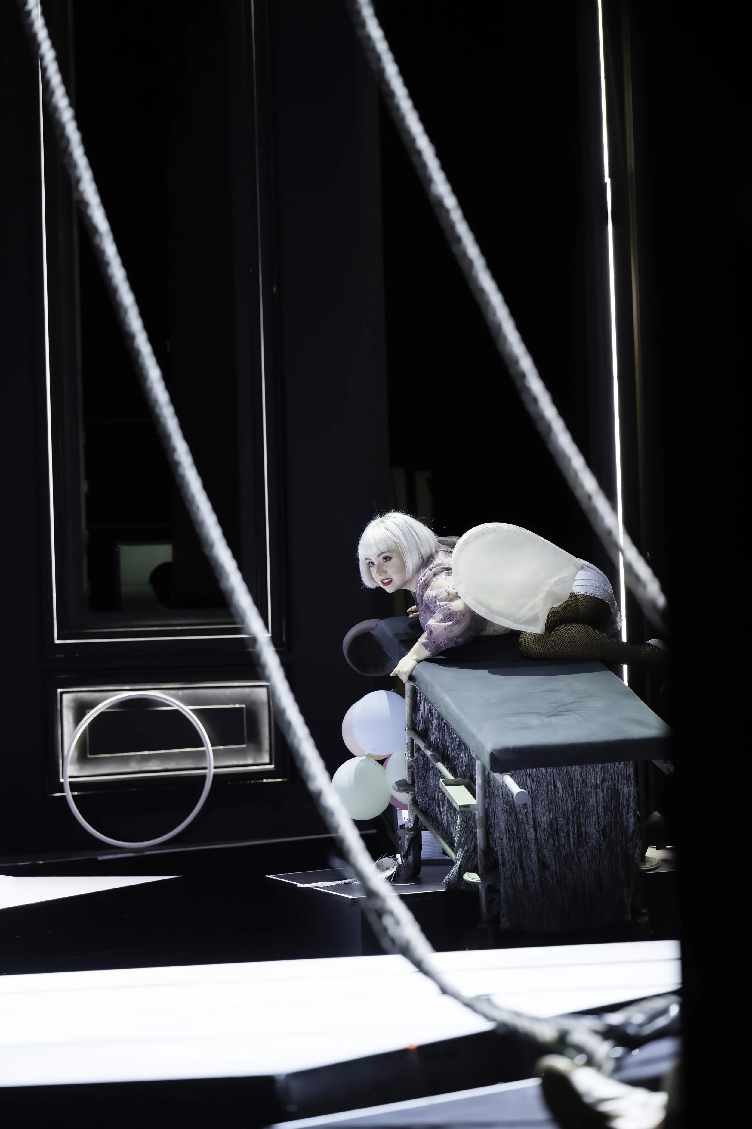A performer with platinum blonde hair crouching on a black stage, dressed in a costume with pink and purple tones, and surrounded by colorful balloons.