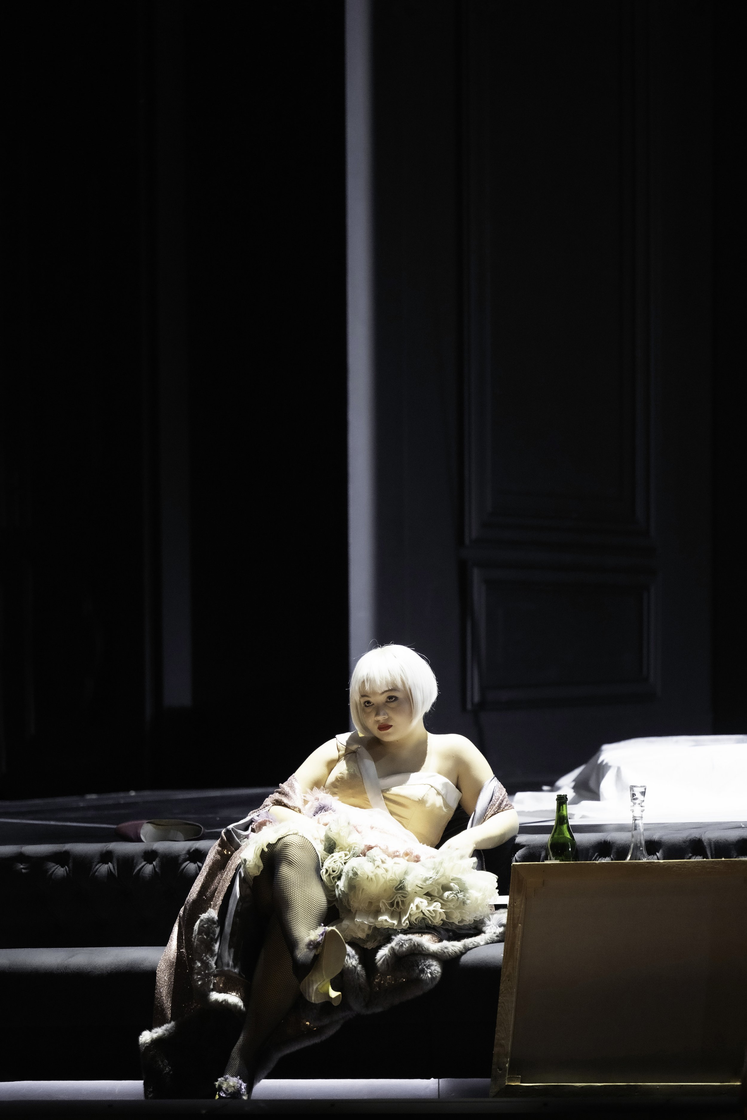 A woman with platinum blonde hair sitting on a stage during a theatrical performance. She is dressed in an elaborate costume with a beige bodice, black fishnet stockings, and a luxurious fur stole. There are bottles and glasses on the table beside he