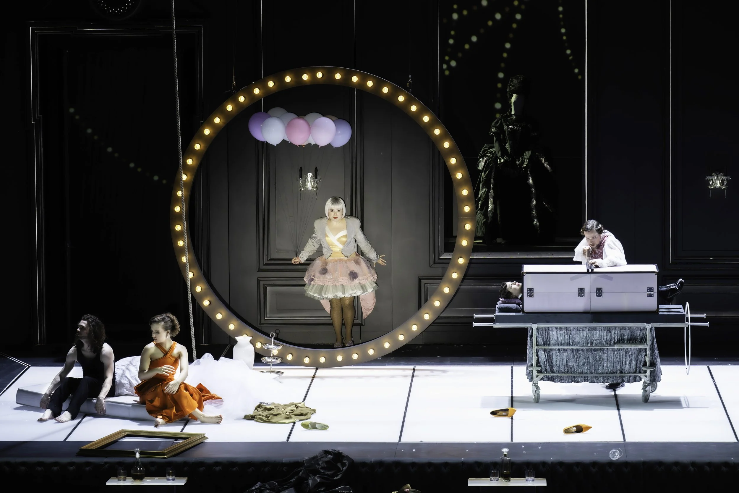 Theatrical stage with performers, one woman in a pink dress with a gray blazer standing inside a large illuminated ring, others sitting or standing around, set with props like balloons, picture frames, and shoes.