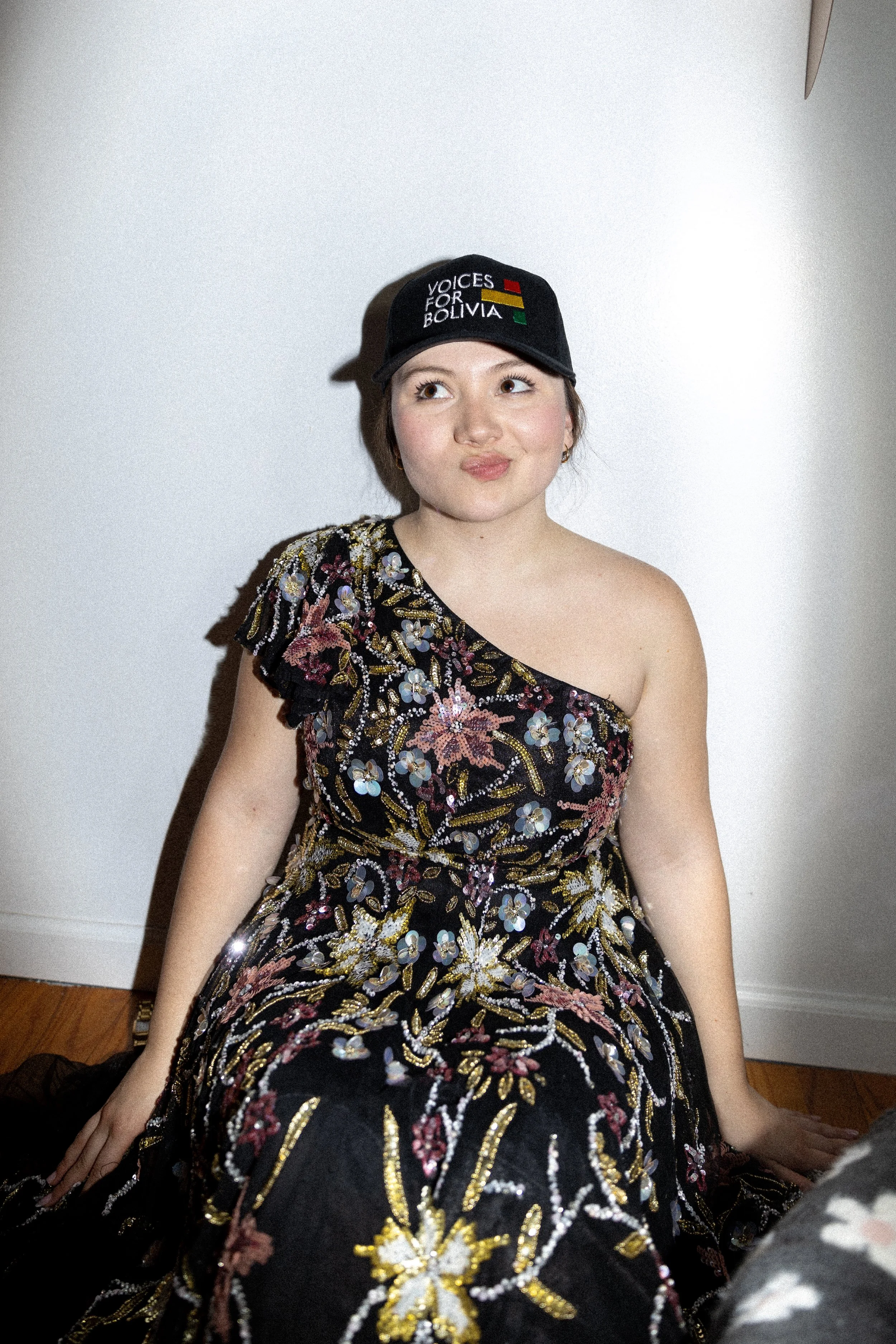 A woman in a floral embroidered dress sitting against a white wall, wearing a black cap that reads 'Voices for Bolivia' with red, yellow, and green flags.