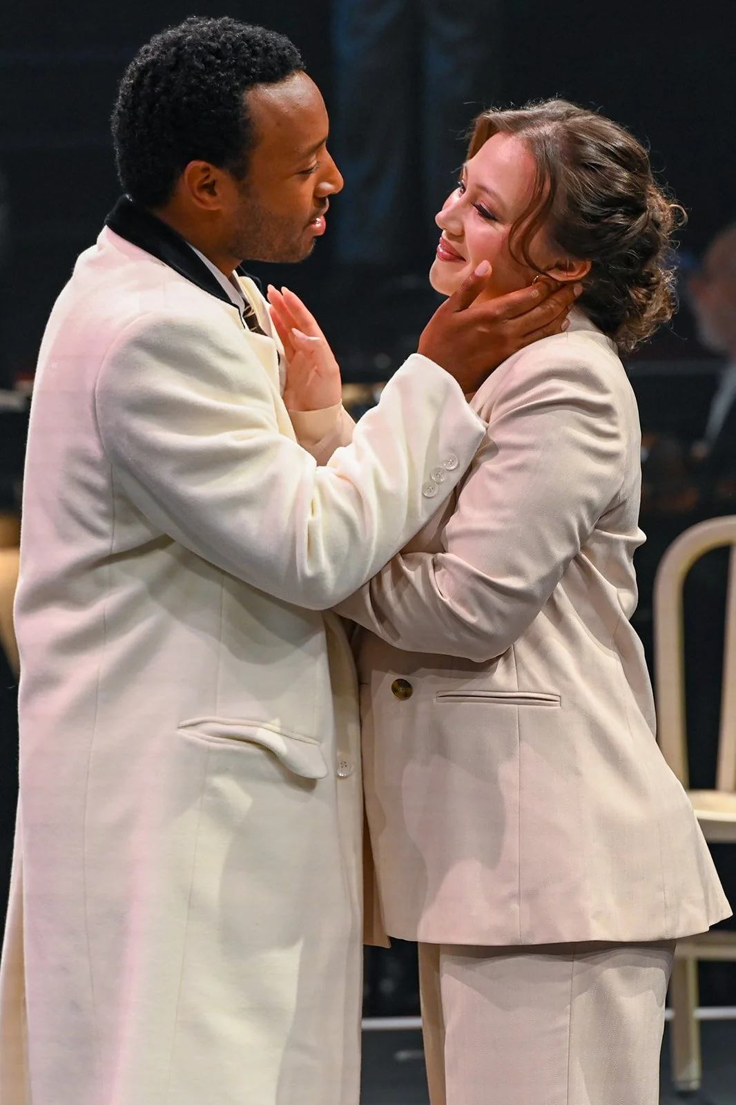 A man and a woman dressed in light-colored suits are sharing an intimate moment, with the man holding the woman's face gently.