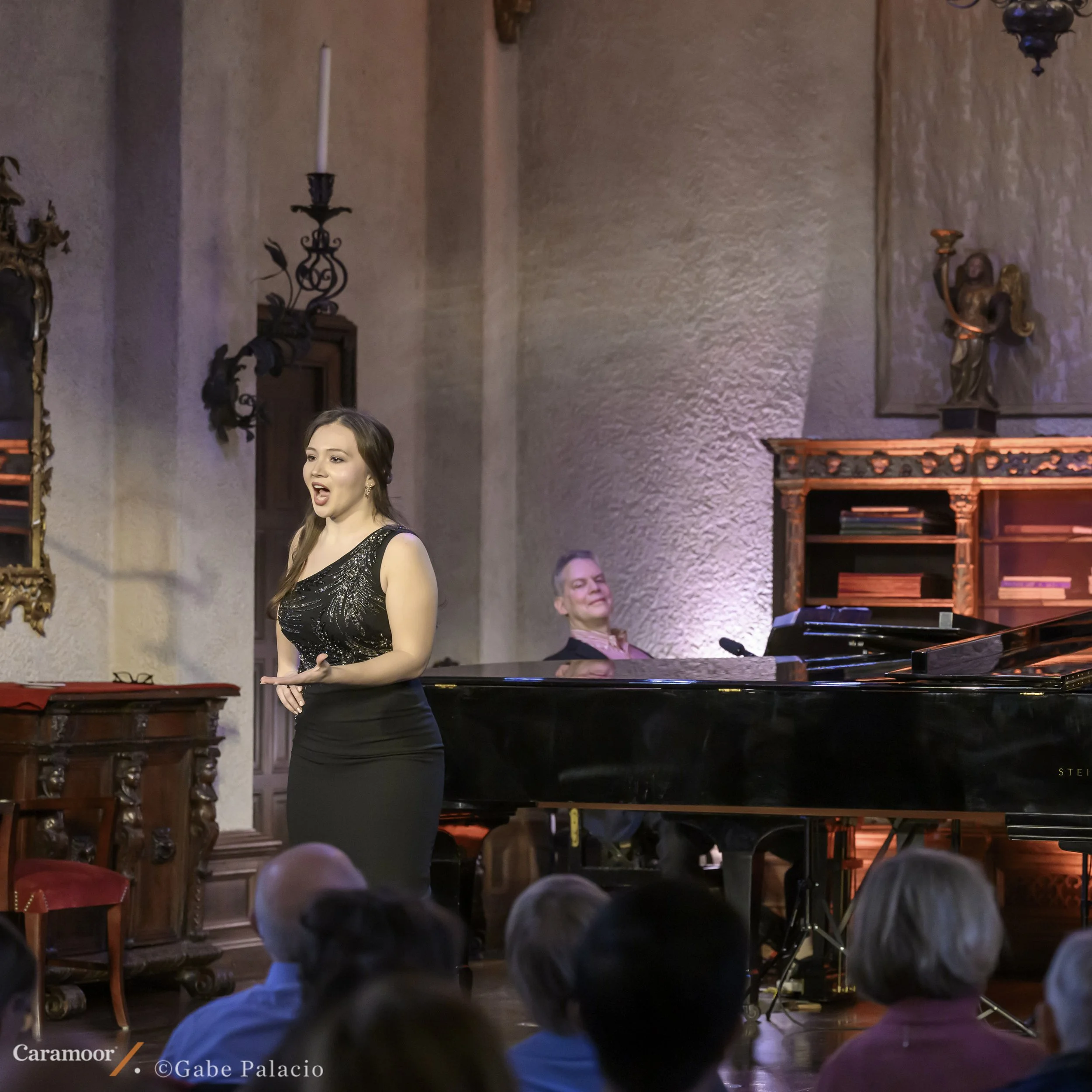 A woman singing at a piano during a performance in a decorated room with ornate wall sconces and statues.