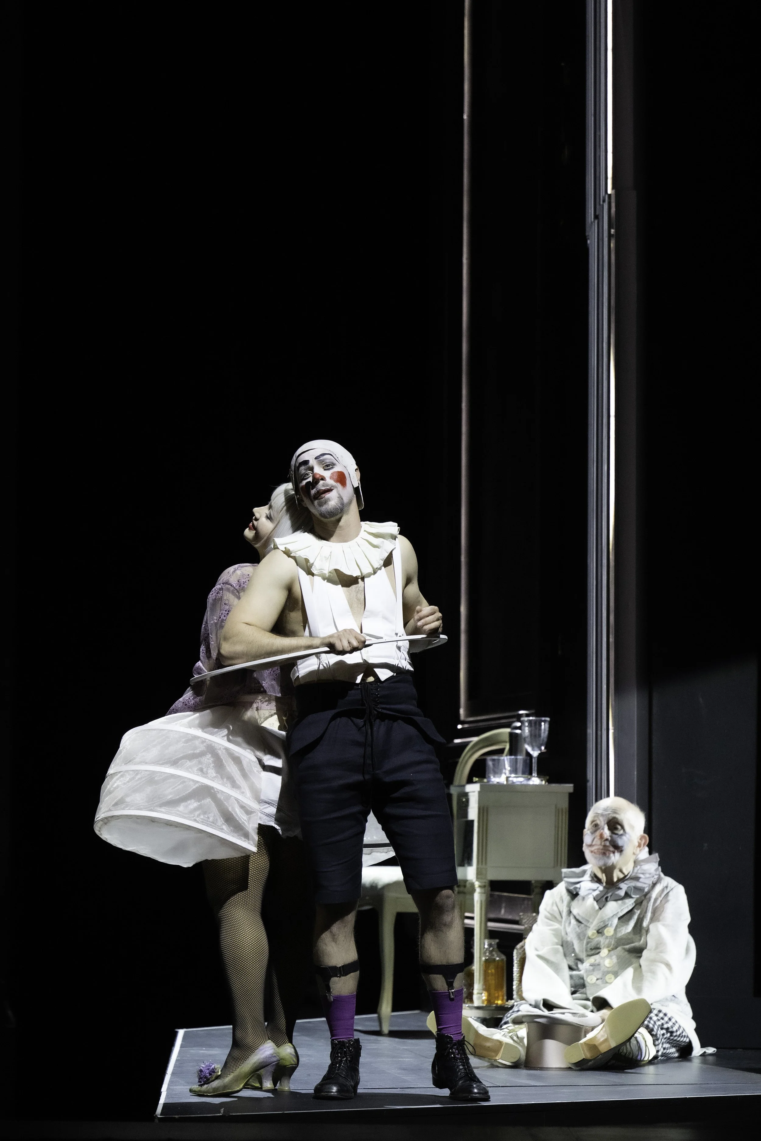 Three performers in theatrical costumes on stage, with black background and minimal set pieces, including a small table and chairs. One performer stands with face painted as a clown, another has a fluffy white dress and makeup, and a third sits on th