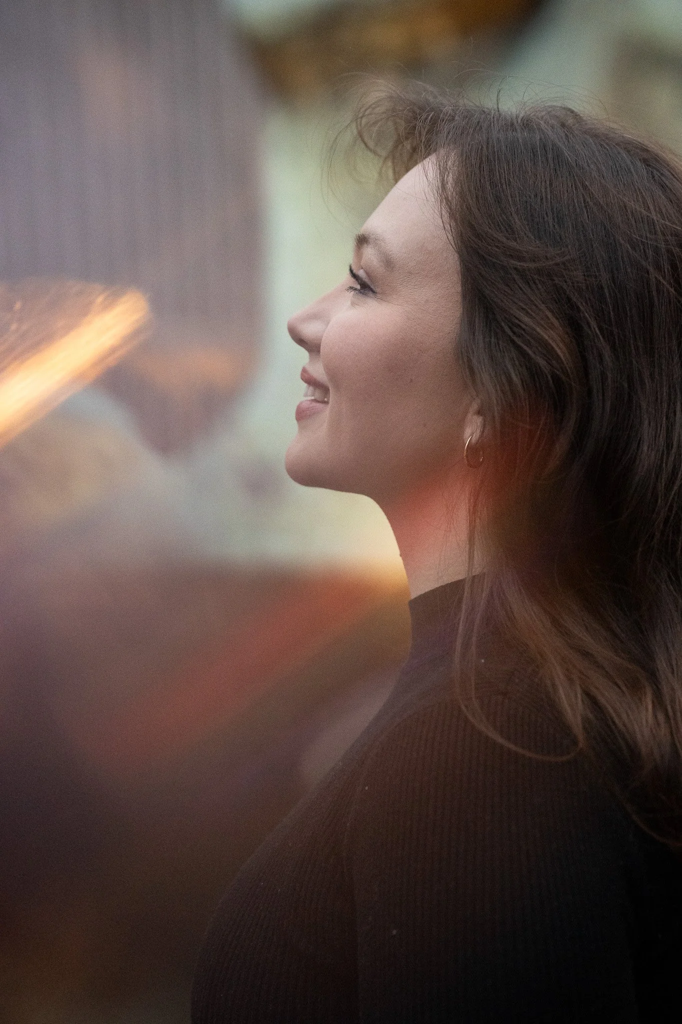 A woman with long brown hair smiling in profile, wearing a dark top and a small hoop earring.