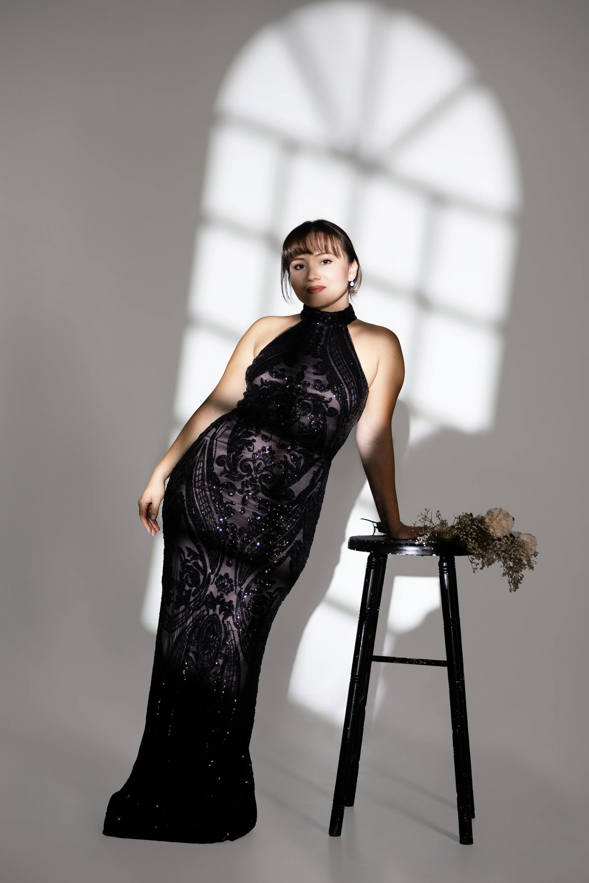 A woman in a black, sheer, sequined dress with intricate patterns stands in front of a large window with an arched top, casting a shadow on the plain wall behind her. A black stool with a bouquet of flowers rests on top of it.