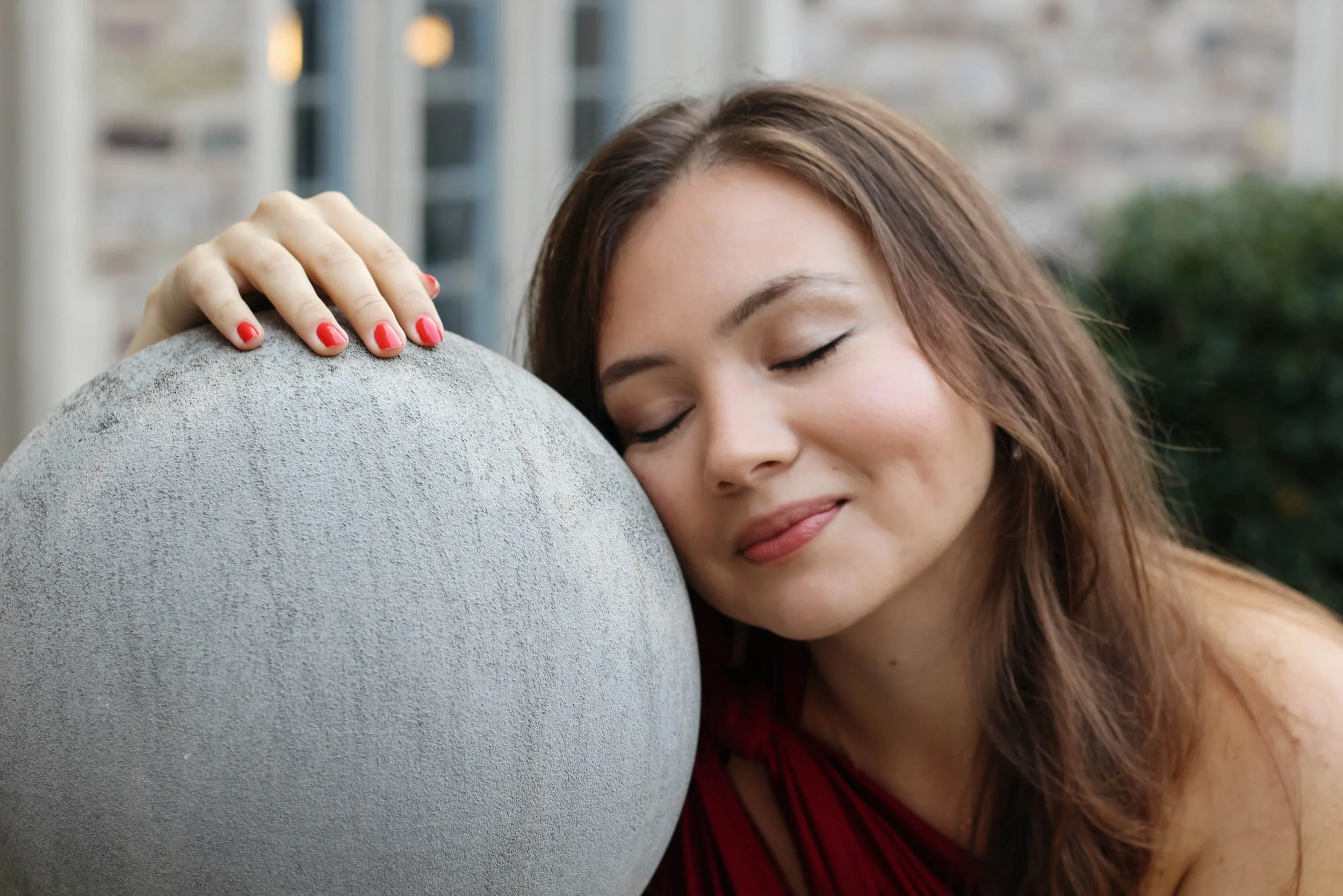 A woman with closed eyes and smiling gently, resting her head against a large, gray concrete sphere during daytime.
