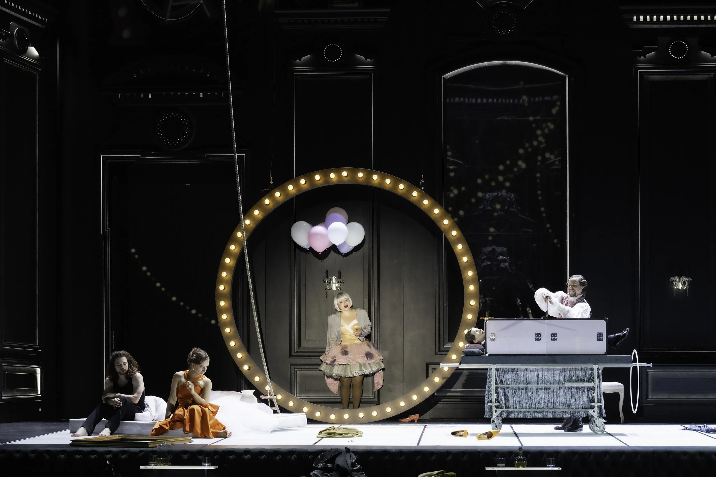 Stage scene with performers and set pieces, including a large circular mirror with lights, a woman dressed in pink with a tutu and black stockings, a woman in orange, and individuals in casual and costume attire, some sitting and others standing, wit