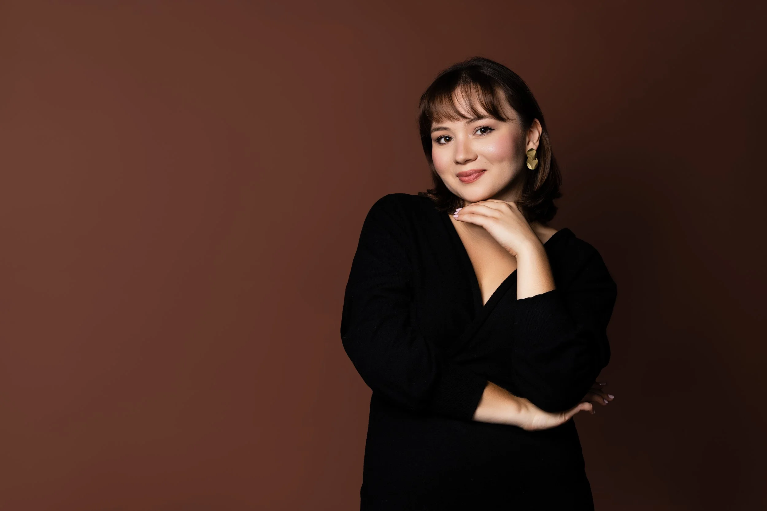 A woman with short dark hair and gold earrings posing with her hand under her chin against a brown background.