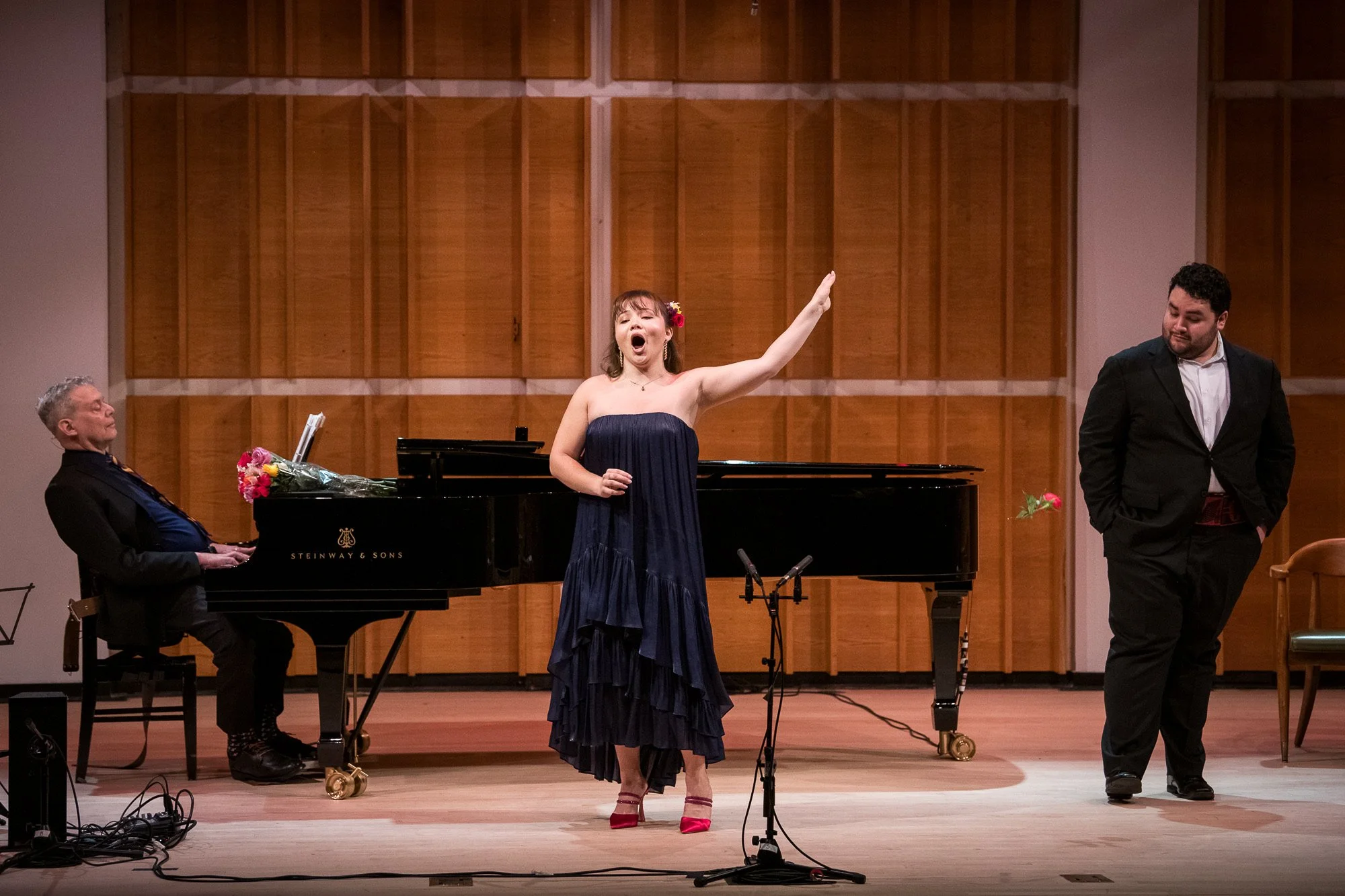 A woman in a strapless dark blue dress and red high heels singing on stage with her right arm raised, a man in a dark suit standing nearby, a man playing a grand piano with flowers on top, and another man dressed in dark clothing in the background.