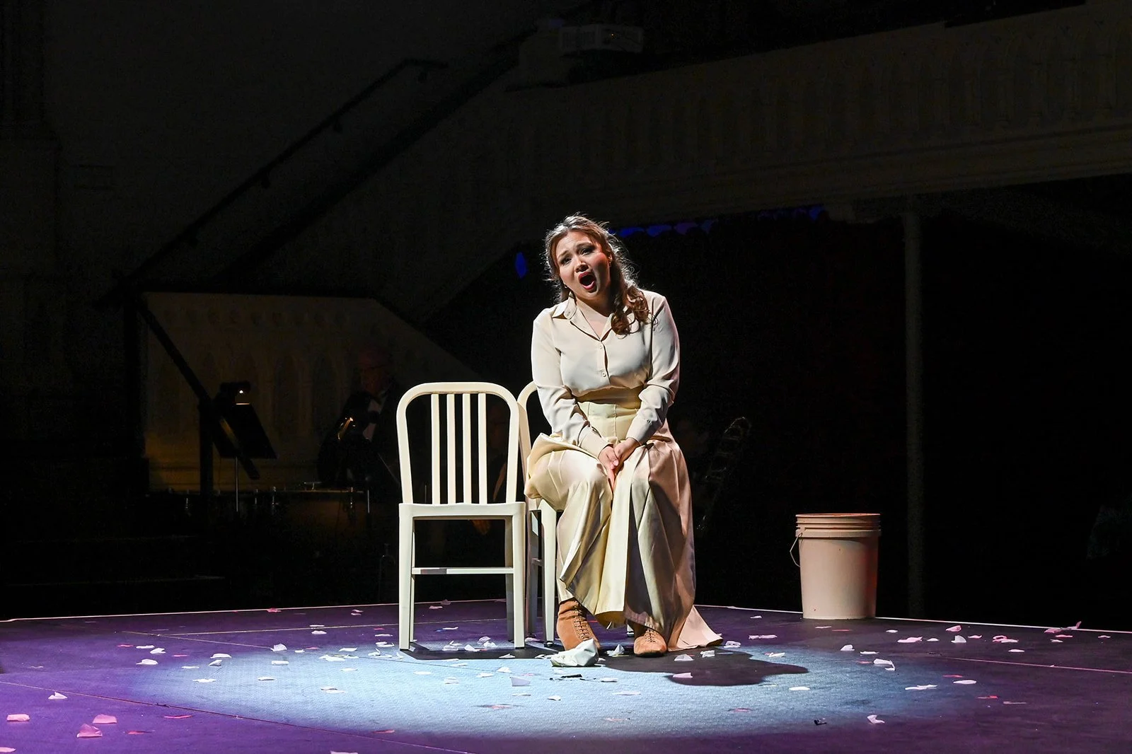 A woman sitting on a white chair on stage, singing or reciting with an expressive face, wearing a cream-colored shirt and long cream pants, surrounded by scattered flower petals and a bucket in the background, with dark surroundings and stage lights 
