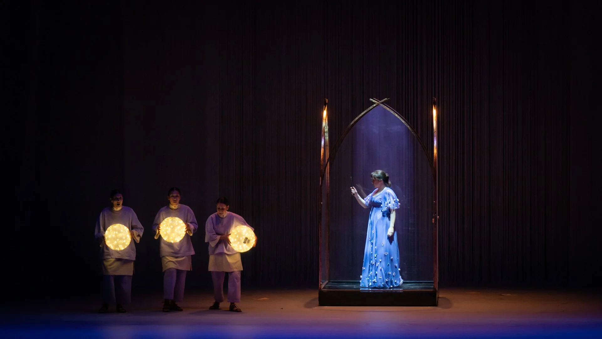 Theater performance with four performers on stage, one woman in a blue dress and three women in gray holding illuminated circular objects, with a dark curtain backdrop and stage lighting.