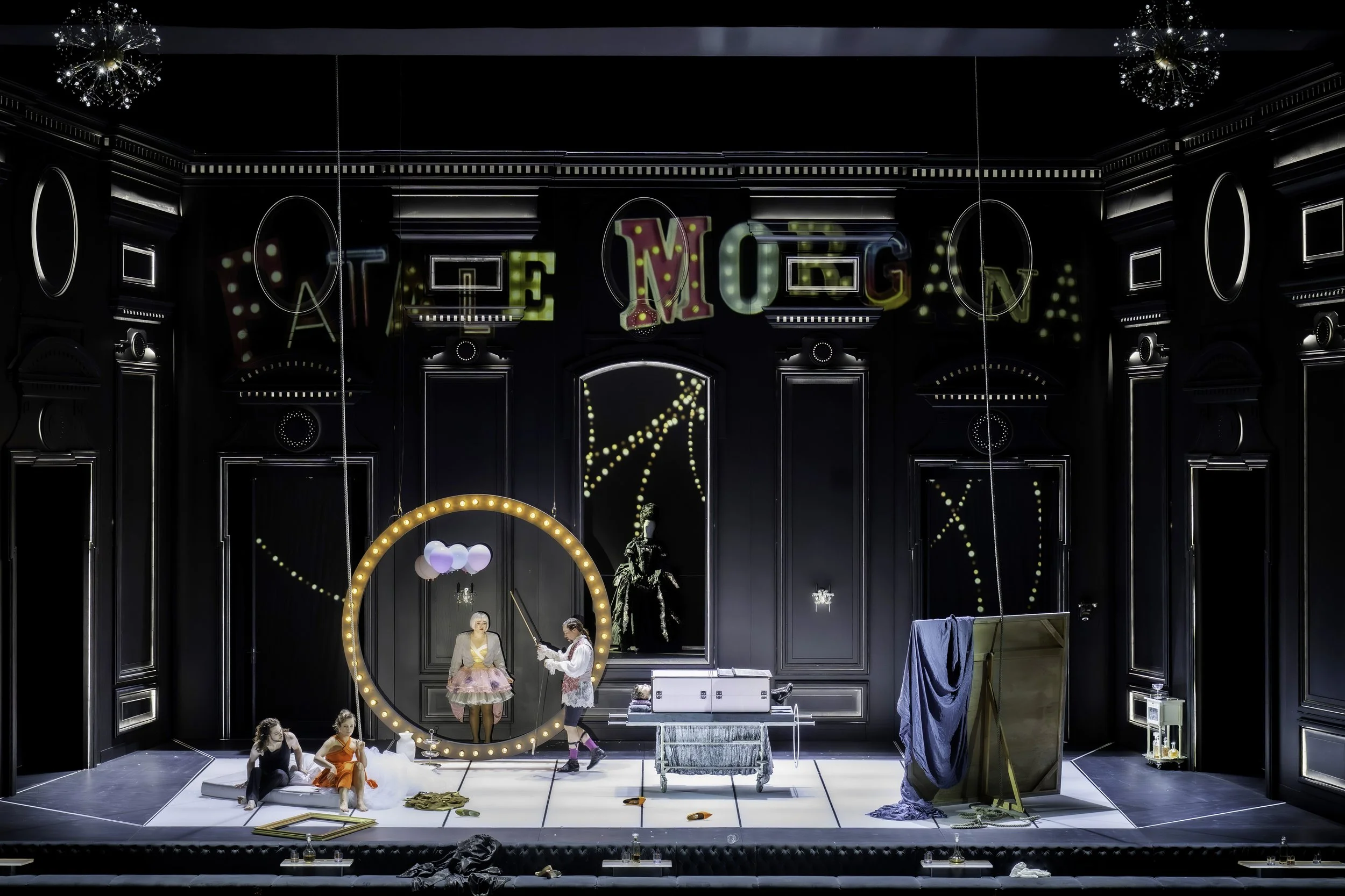 Stage set with black walls and decorative trim, colorful marquee sign spelling 'ARTE YUGO' at the top, and various props like a round mirror with lights, balloons, costumes, and a woman in a pink dress seated on a white platform. Several people are p