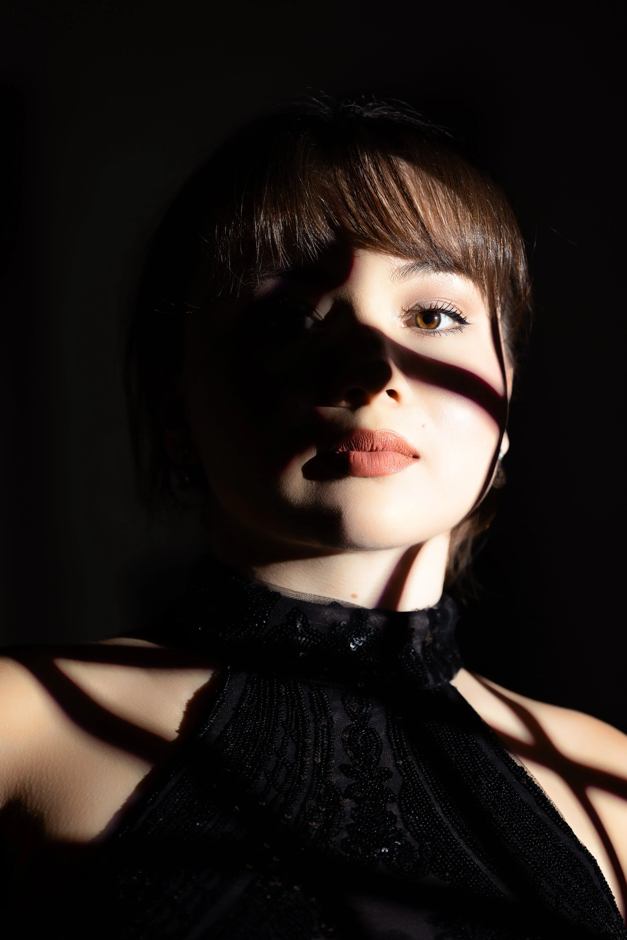 A woman with brown hair and light makeup in a black lace dress, illuminated by a beam of light highlighting her face and shoulder against a dark background.