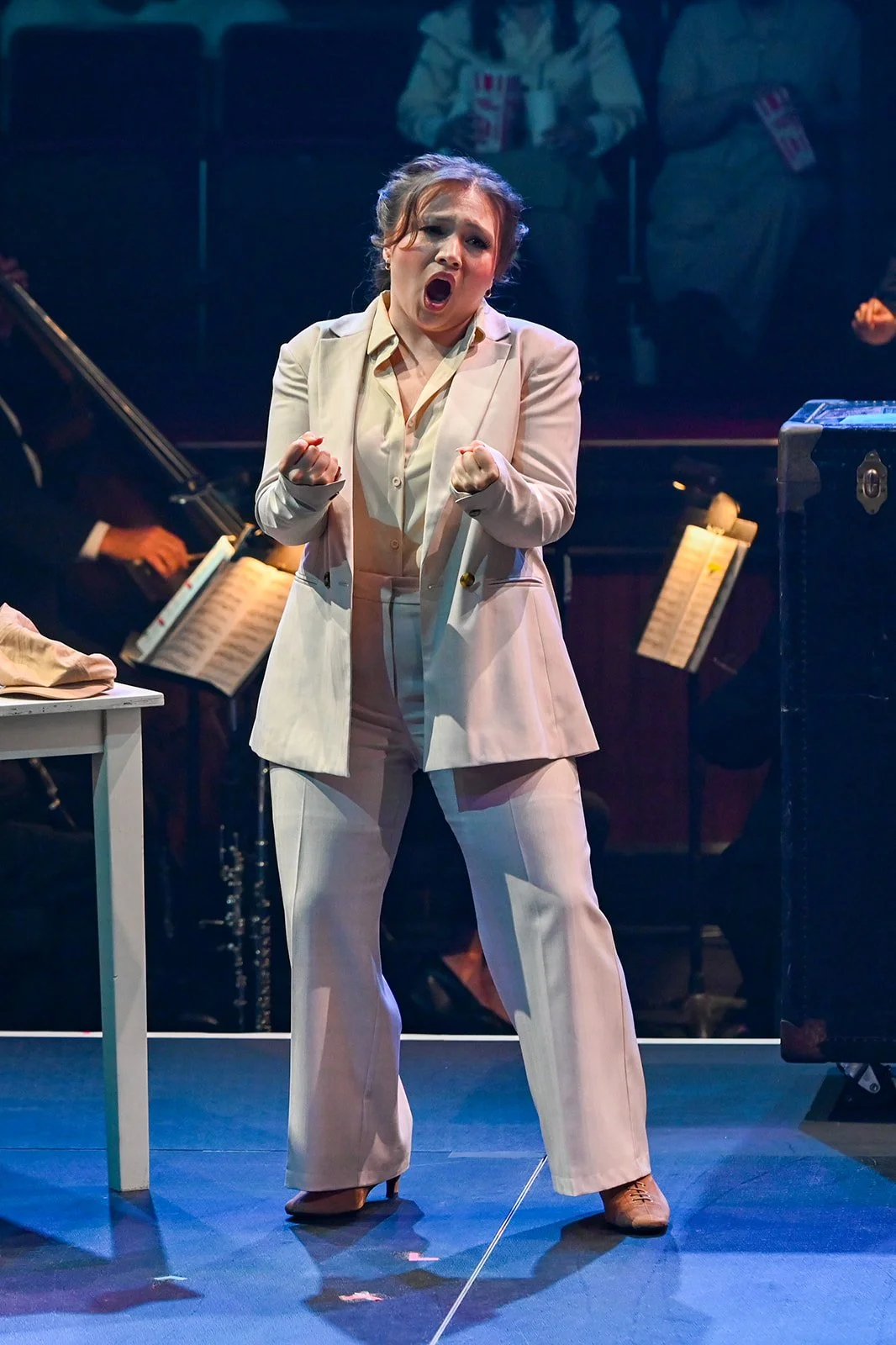 A woman in a cream-colored suit passionately singing on stage during a performance, with music stands and orchestra members in the background.