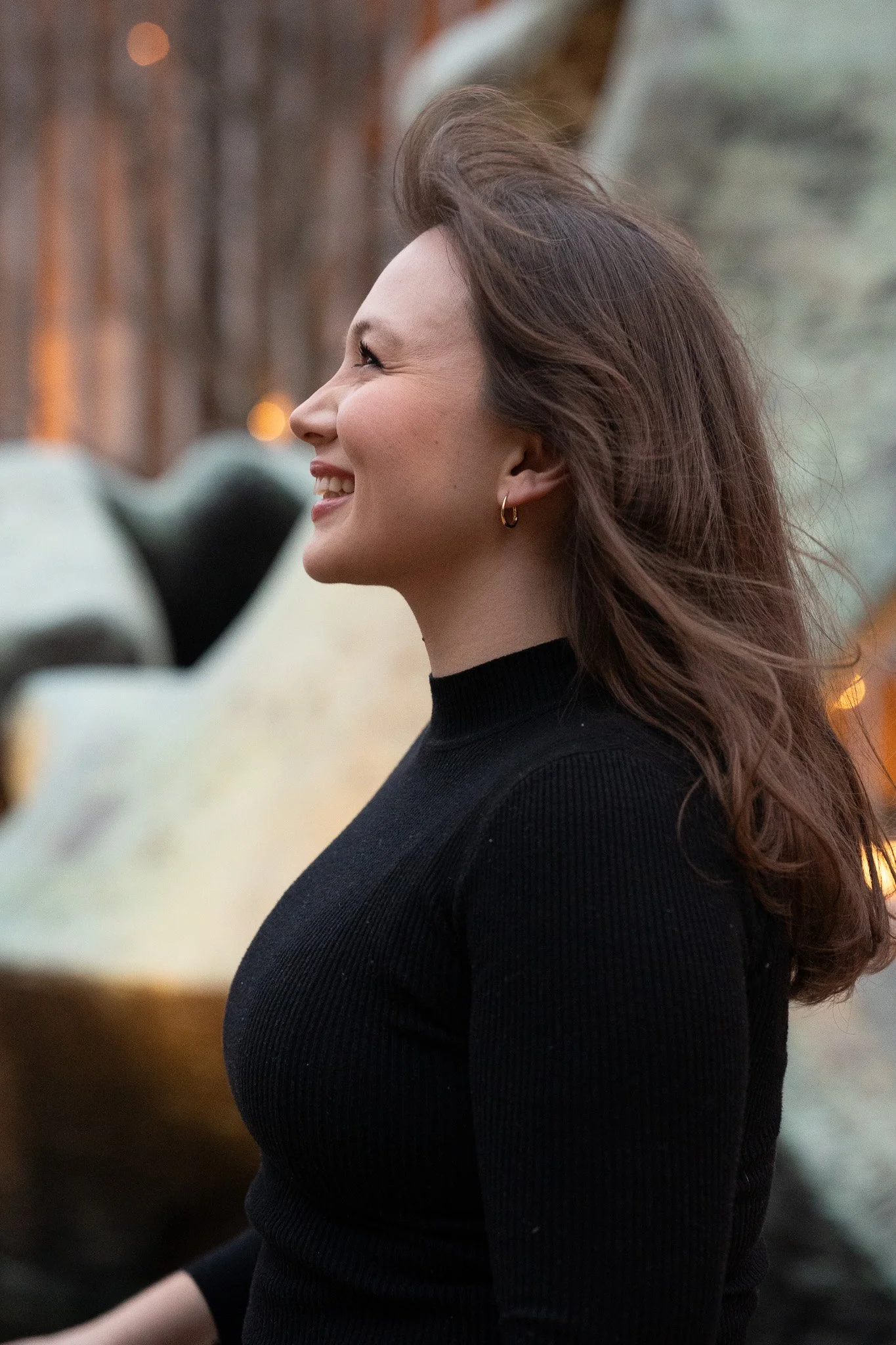 Profile of a smiling woman with long wavy brown hair, wearing a black turtleneck sweater and small hoop earrings, outdoors during fall with blurred background.