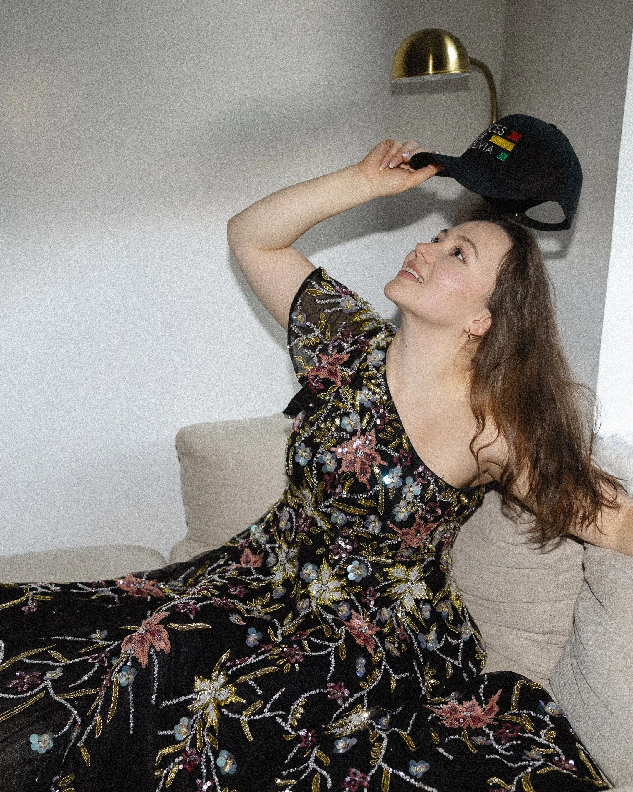 A woman with brown hair smiling and looking up, wearing a black embroidered dress with floral patterns and holding a black cap with medals and flags logo, sitting on a beige couch against a plain wall with a gold wall lamp.