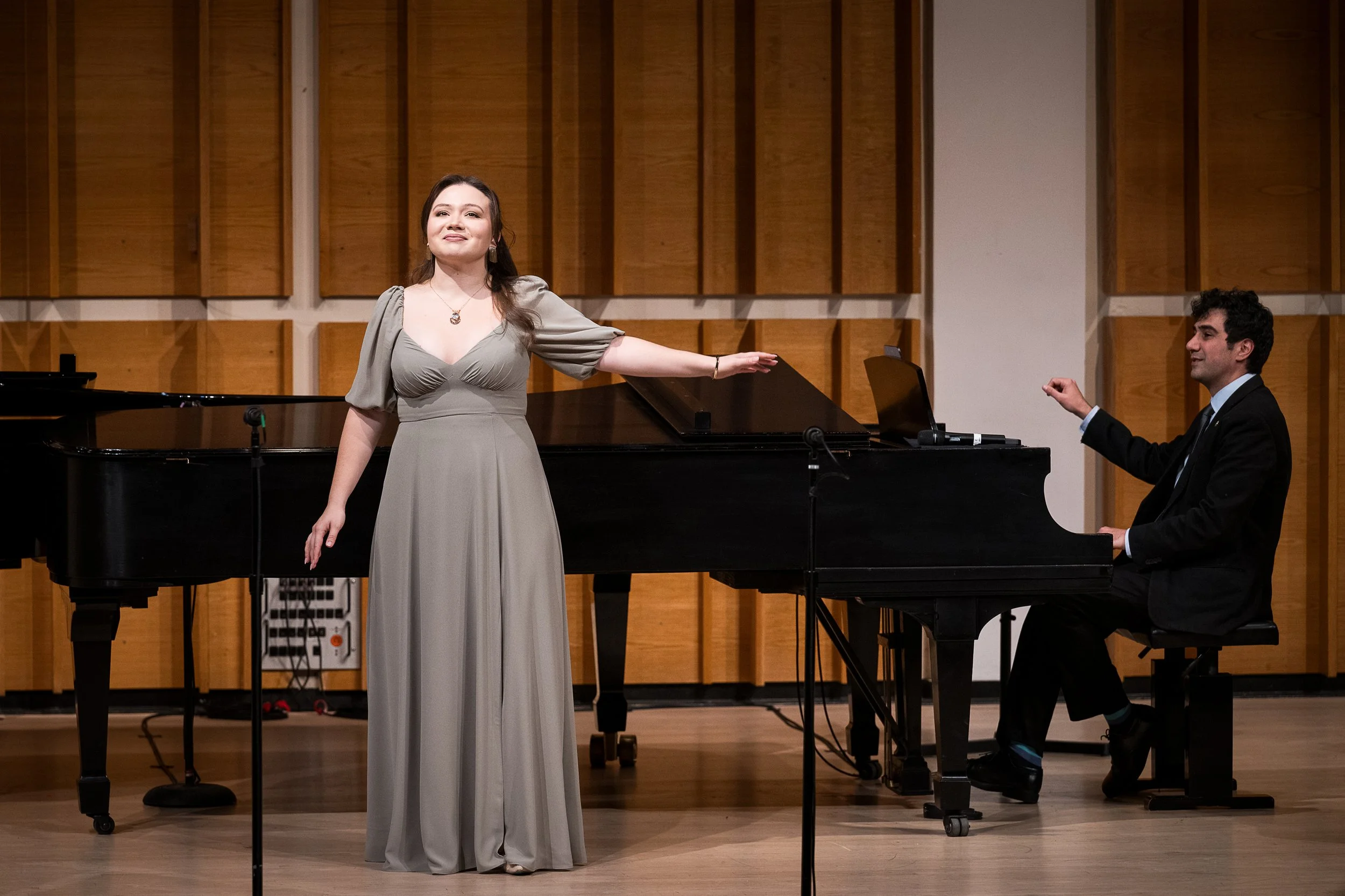 A woman in a long gray dress standing at a microphone with her arms extended, in front of a grand piano, in a room with wooden acoustic panels and a man seated at the piano in a black suit, playing.