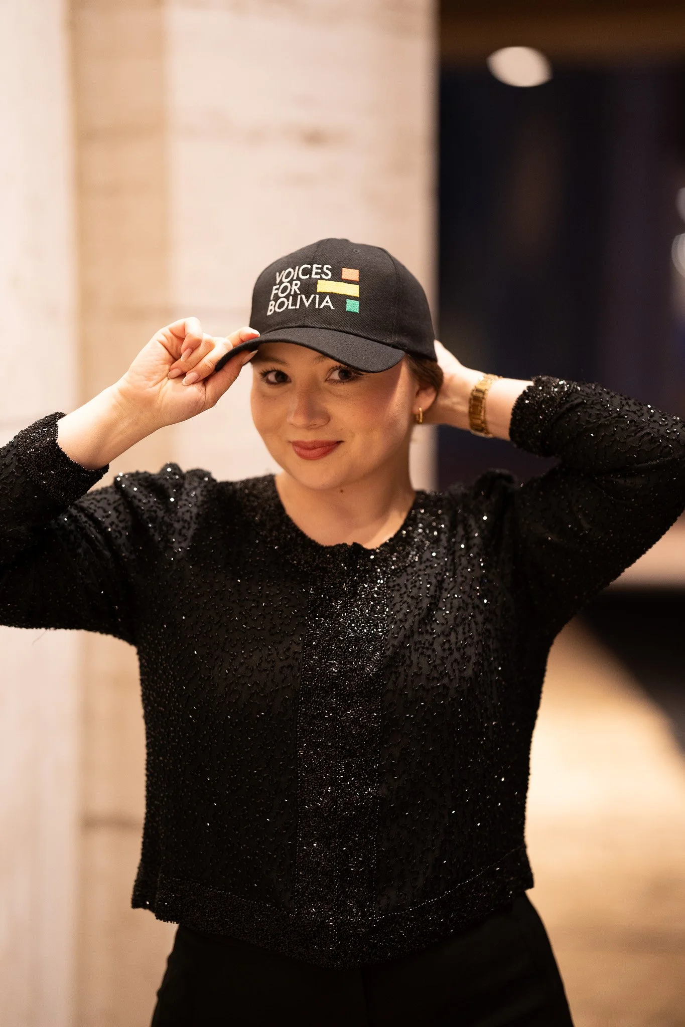 A woman in a black, sparkly long-sleeve top adjusting a black baseball cap with 'Voices for Bolivia' and a flag design.