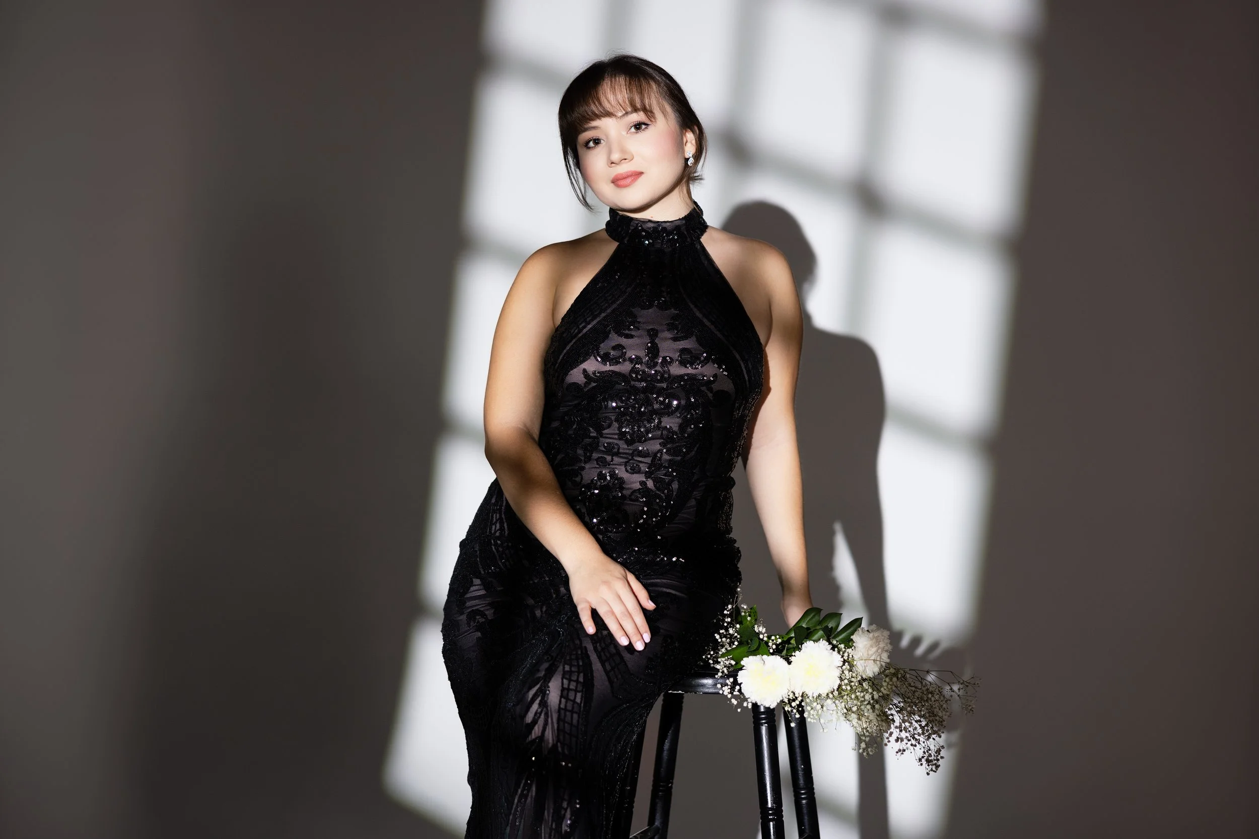 A woman in a black dress sitting on a stool next to a bouquet of white flowers, with light coming through a window casting shadows.