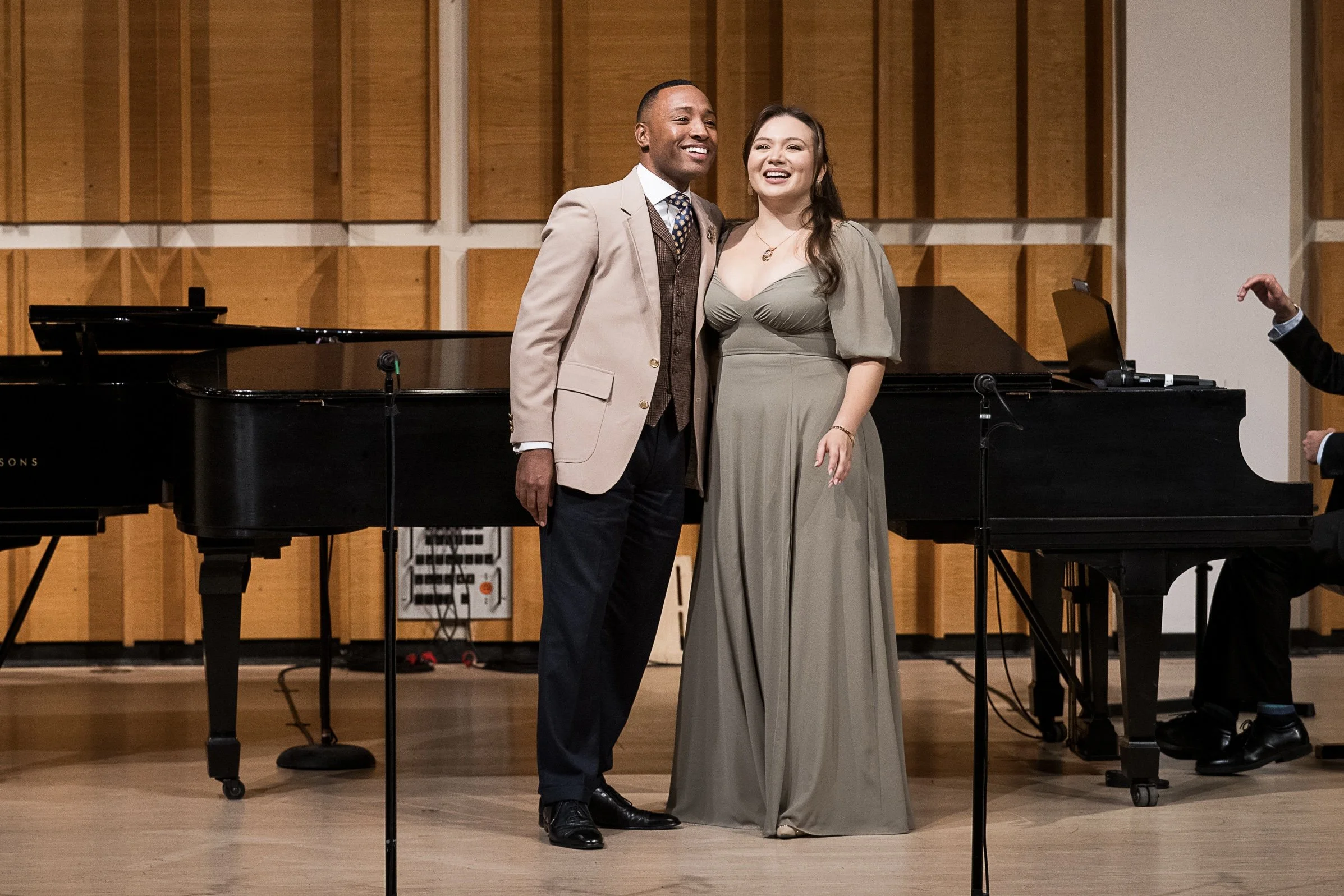 A man and woman smiling and posing on stage after a performance, standing in front of a grand piano, with wood paneled soundproofing on the wall behind them.