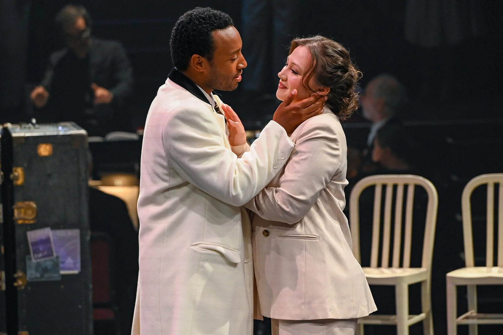 A man in a white suit gently holds a woman’s face and neck as they gaze into each other's eyes during a performance on stage.