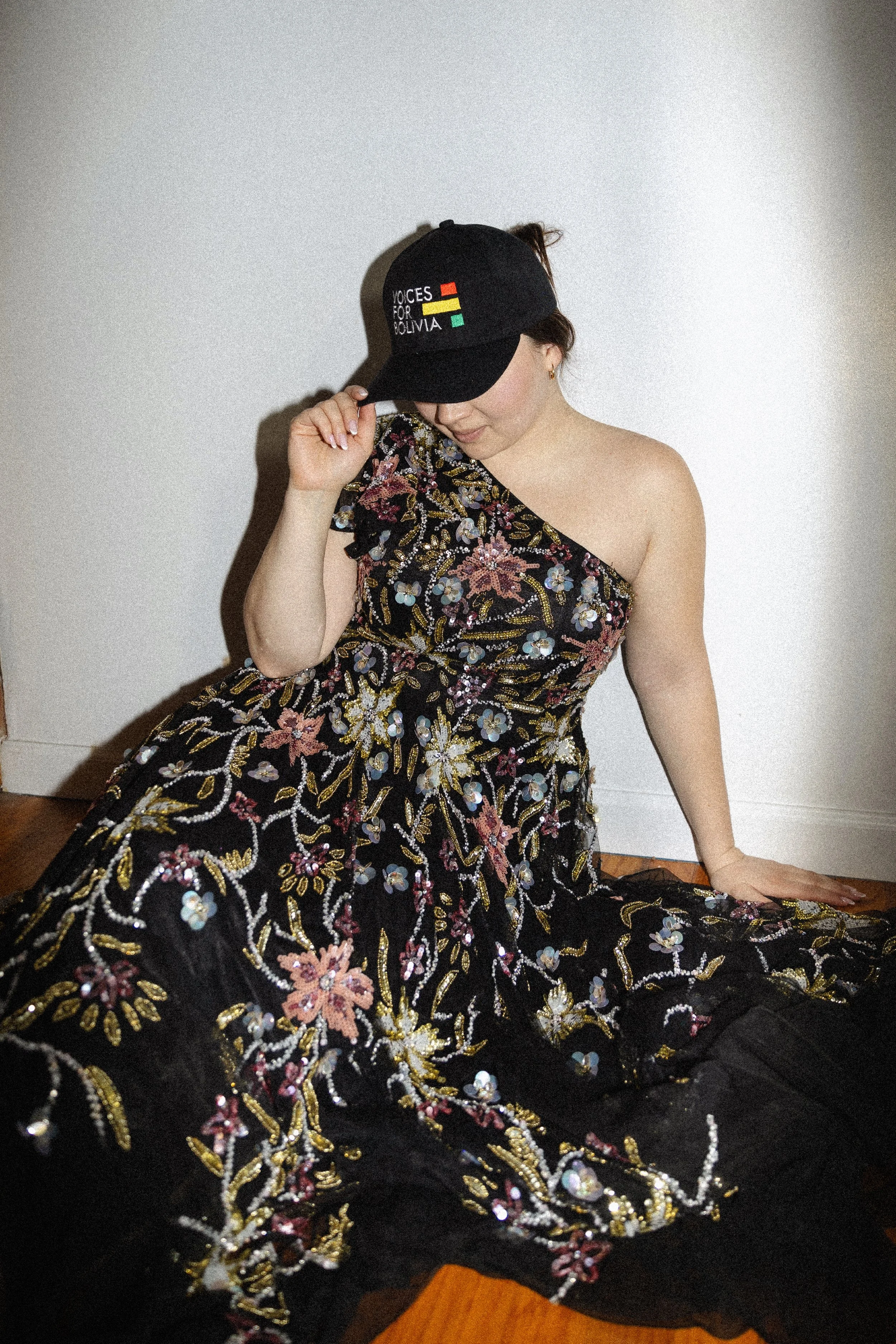 A woman in a strapless floral dress sitting on the floor, holding a black cap with the words "VOICES FOR BOLIVIA" and colored flags. The background is a plain white wall.