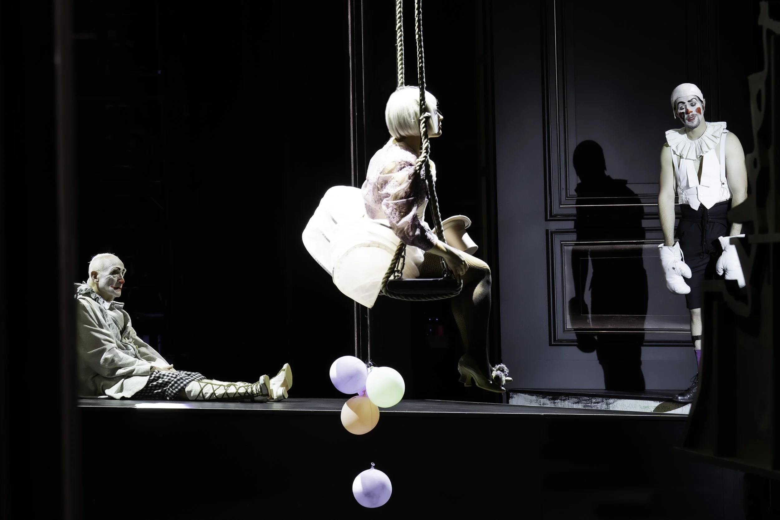 Clowns performing on stage with one clown sitting on a swing, another clown standing with white gloves, and a third clown lying on the floor, all dressed in theatrical costumes and makeup.