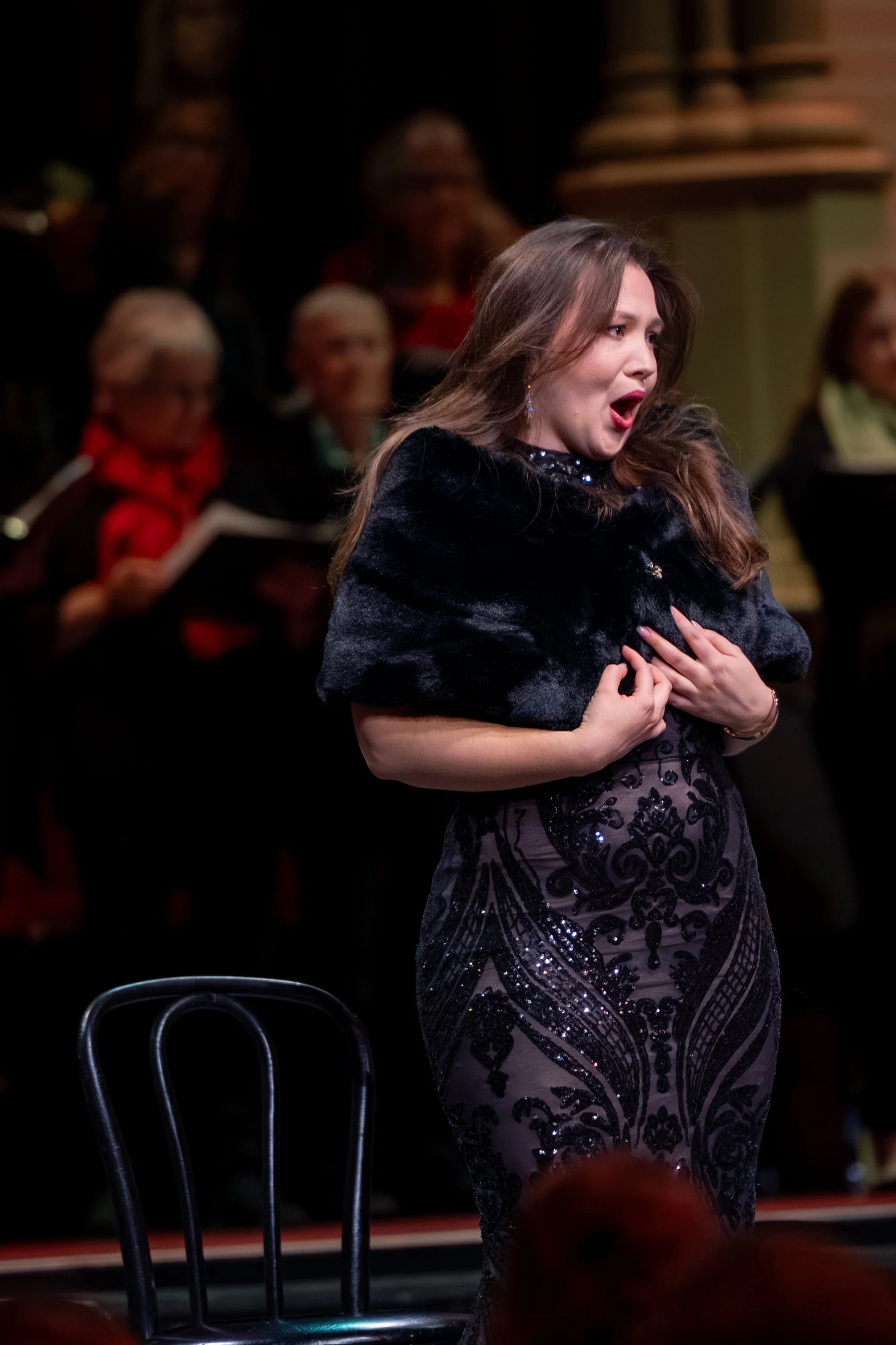 A woman with long brown hair wearing a dark, sparkly dress is singing passionately on a stage, with her hands crossed over her chest. Behind her, a group of people, mostly women, are seated and watching her performance.