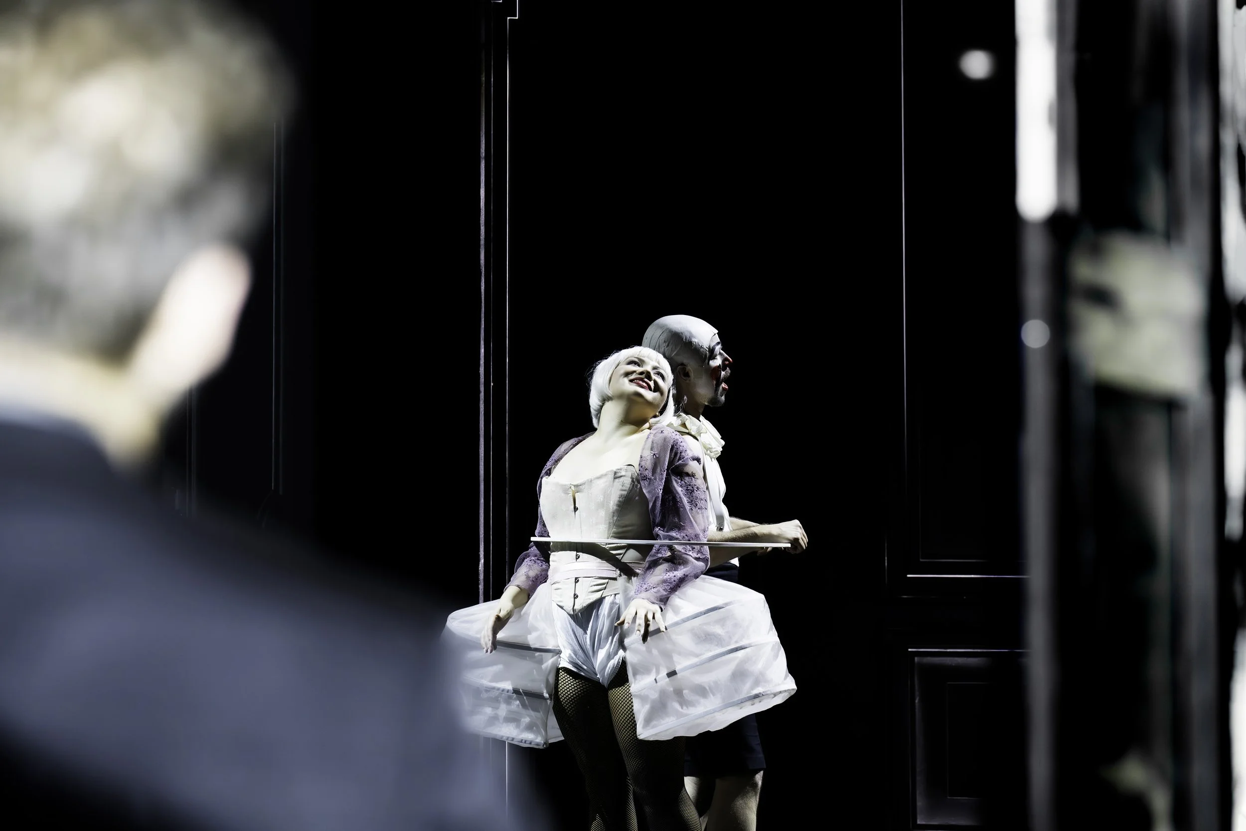 Two performers in vintage costumes sitting in an elevator with mirrored walls, one smiling and the other looking away.