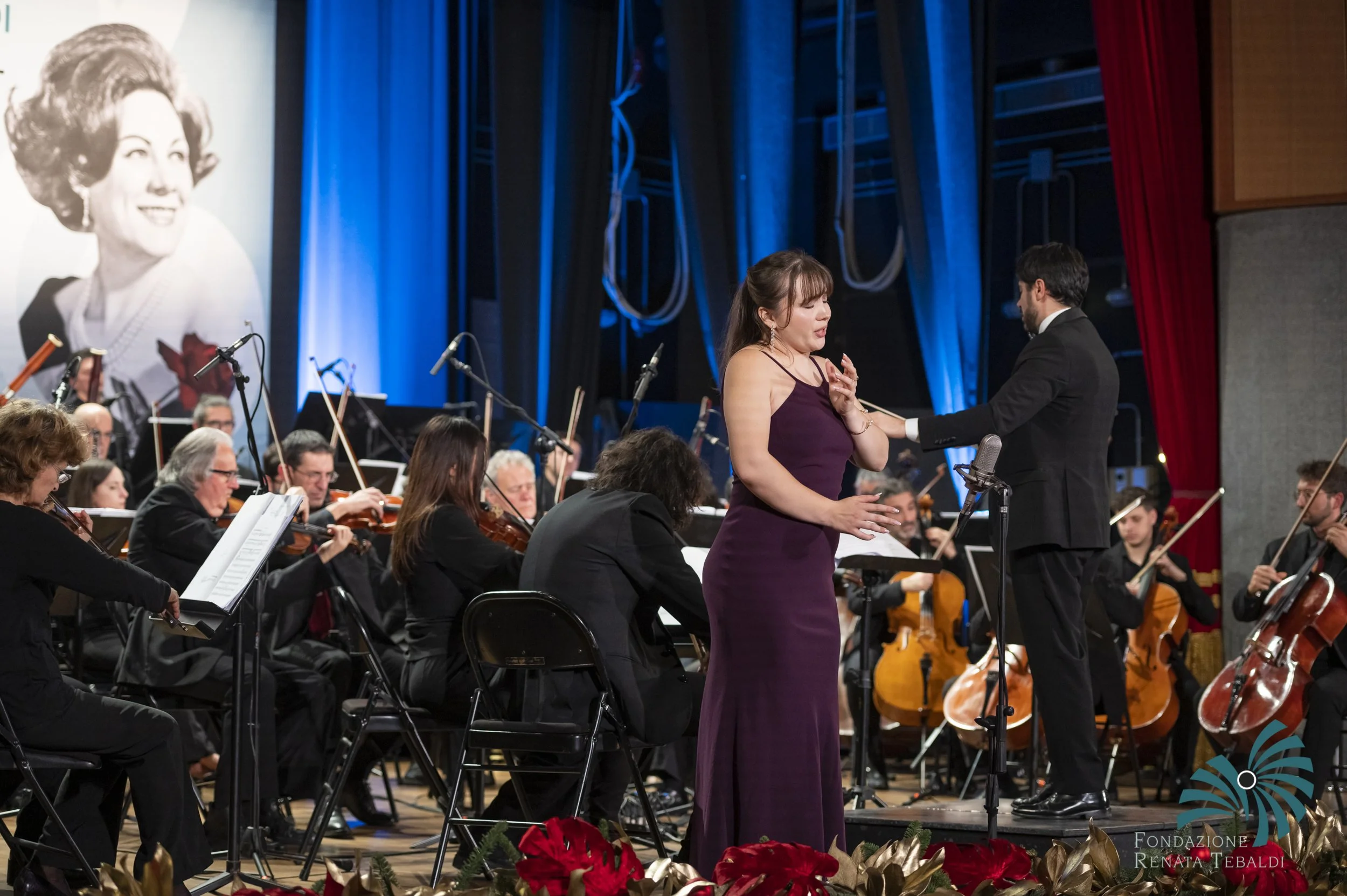 A woman in a purple dress singing on stage during an orchestral performance, with a conductor in a suit behind her, and orchestra members playing instruments in the background, decorated with red flowers and green foliage.