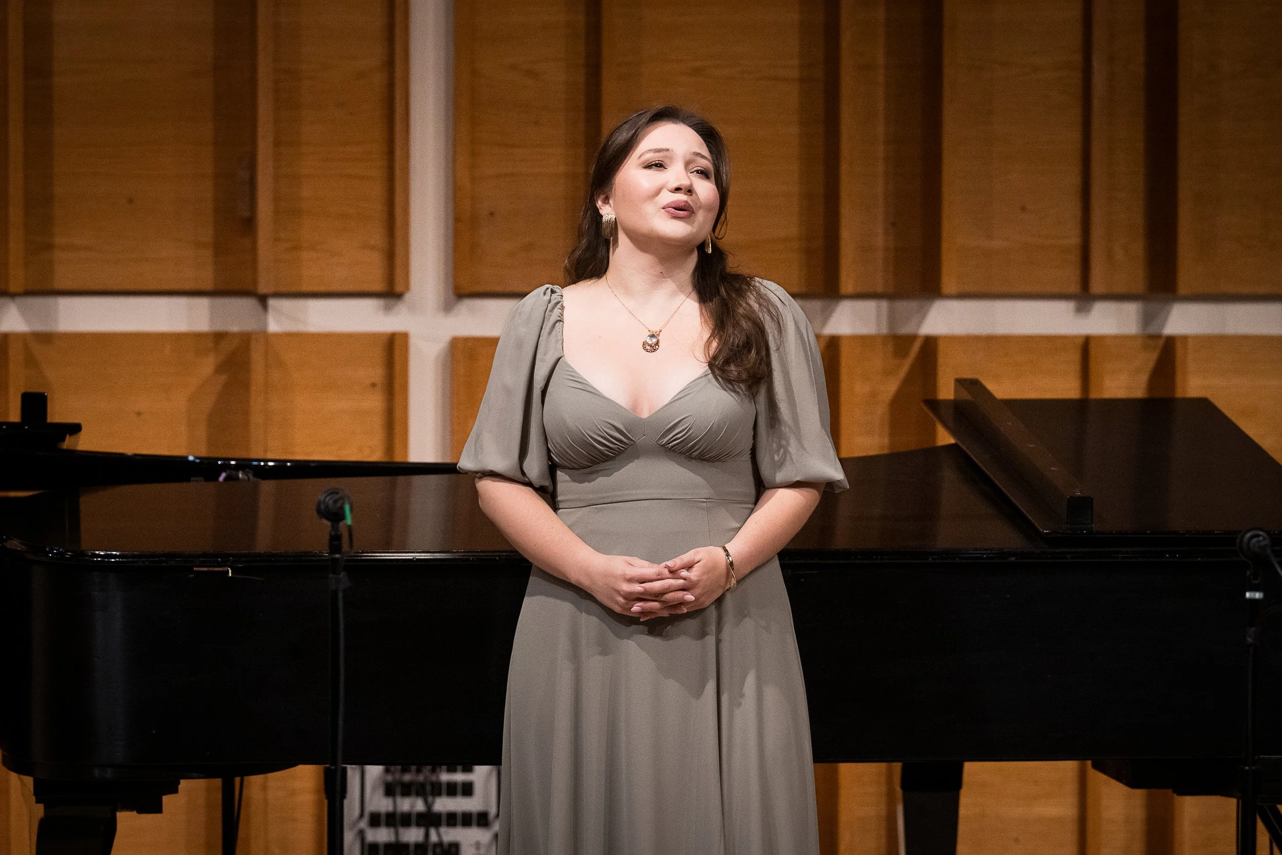 A woman standing in front of a grand piano in a music studio, surrounded by wooden acoustic panels.