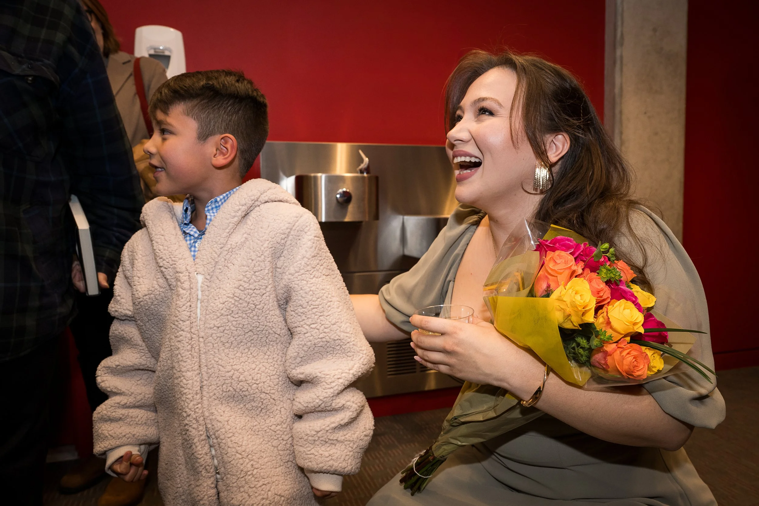 A woman with long brown hair, wearing earrings and a beige top, is smiling and holding a bouquet of colorful roses and a glass of drink. She appears to be greeting or congratulating a young boy with short dark hair, who is wearing a beige fleece jack