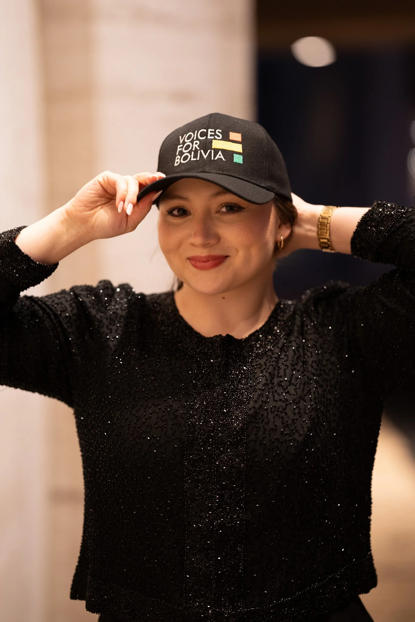 A woman wearing a black sparkly top and a black baseball cap with the words 'Voices for Bolivia' and colorful bars. She is smiling and adjusting her cap with both hands, wearing a gold watch and small hoop earrings.