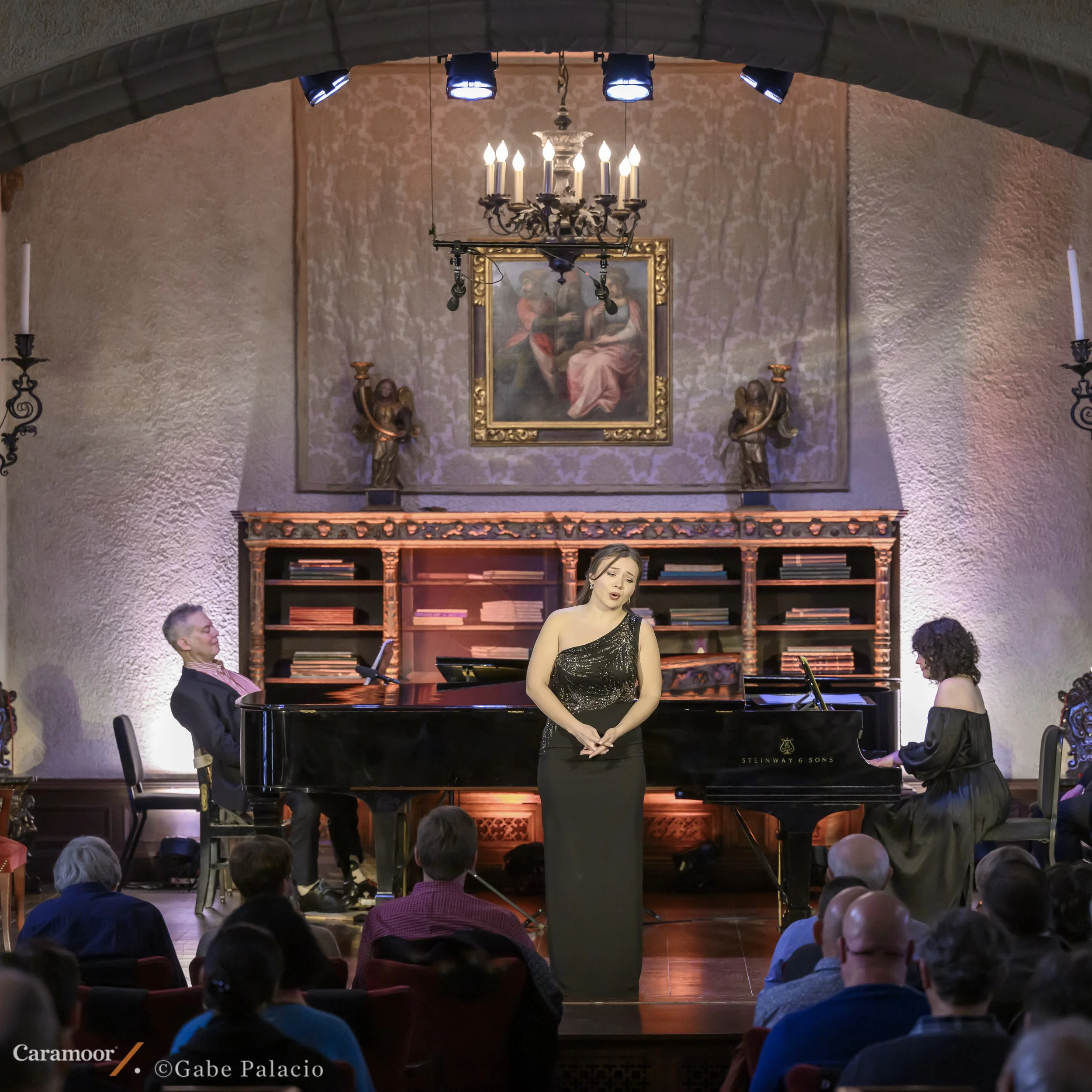 A woman performing at a piano concert in front of an audience, with a chandelier hanging from the ceiling and a painting of two women behind her.