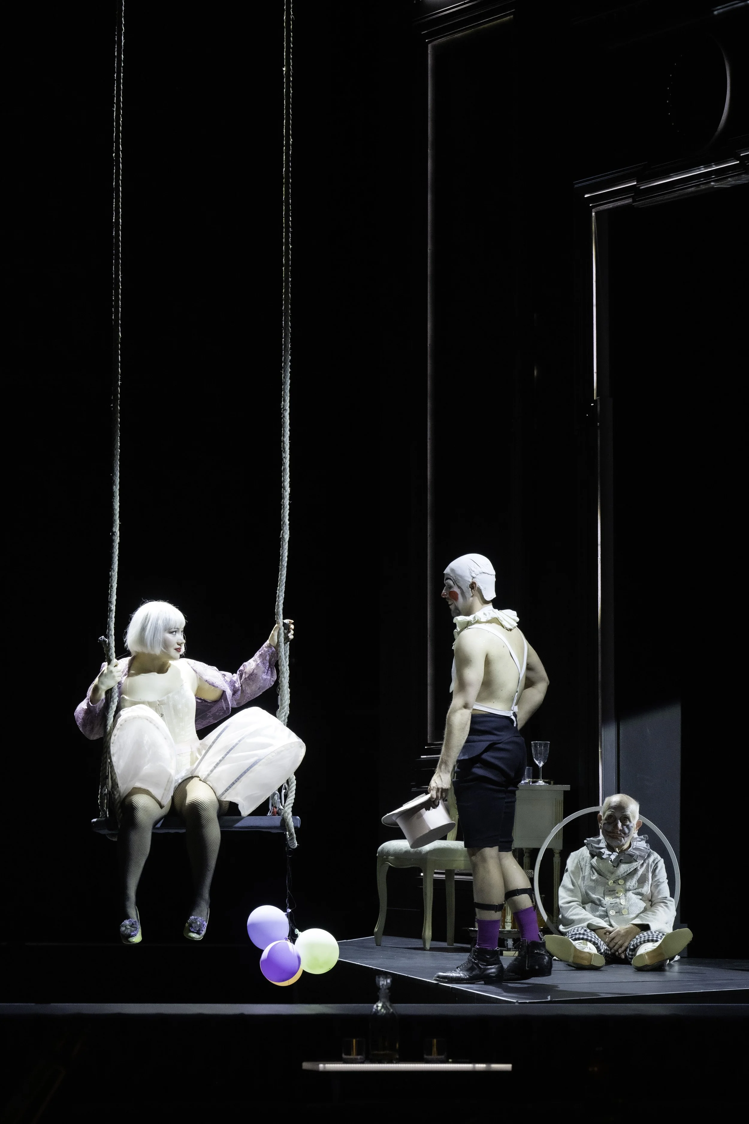 Stage scene with five performers dressed in vintage costumes. Two are seated on a swing, one in a purple dress with a short blond bob, and another in a white dress. Two performers are standing; one male with a bare chest, shorts, and purple socks, ho