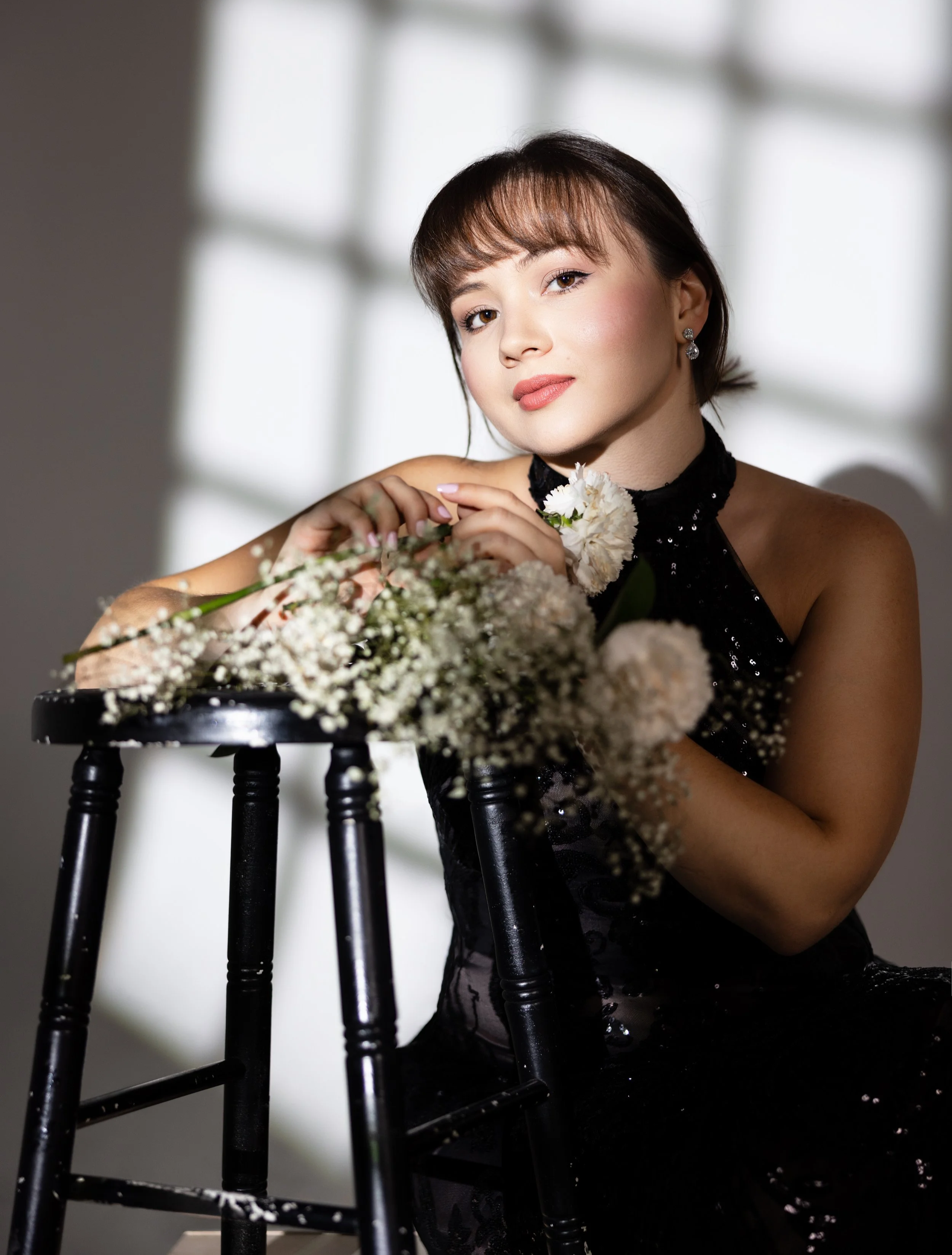 A woman with short brown hair and earrings, dressed in a black sequined dress, sits behind a black wooden stool with floral arrangements on it, in a softly lit room with a bright window in the background.