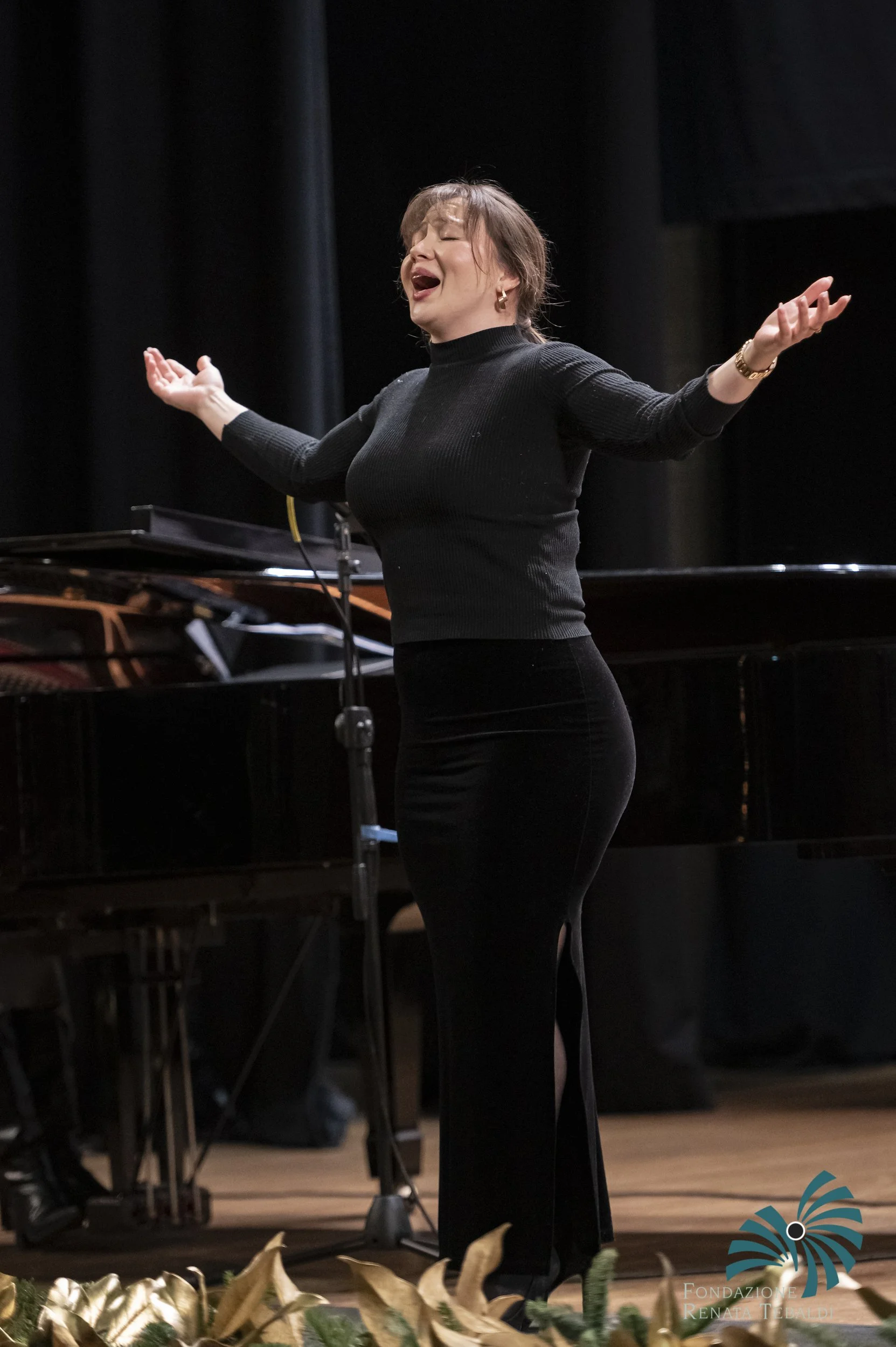 A woman passionately singing on stage with her eyes closed, wearing a black long-sleeve top and earrings, standing in front of a grand piano, with black curtains in the background and floral decorations at the front of the stage.