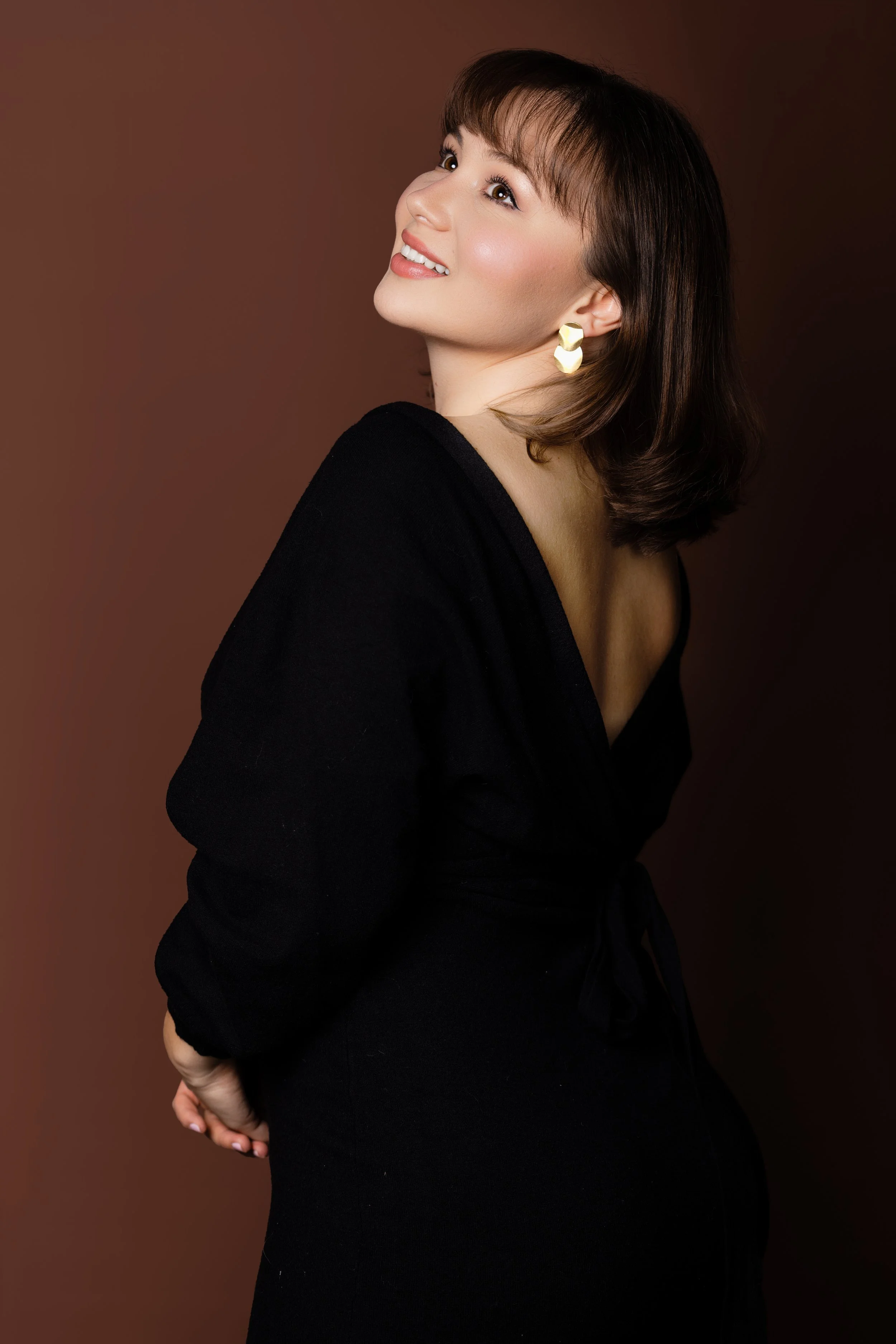 A woman with short dark hair and earrings, smiling and looking up, wearing a black top against a brown background.