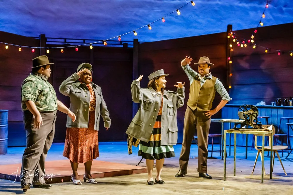 Five actors performing on an outdoor stage at dusk, with three women and two men, during a theatrical play, with string lights overhead.
