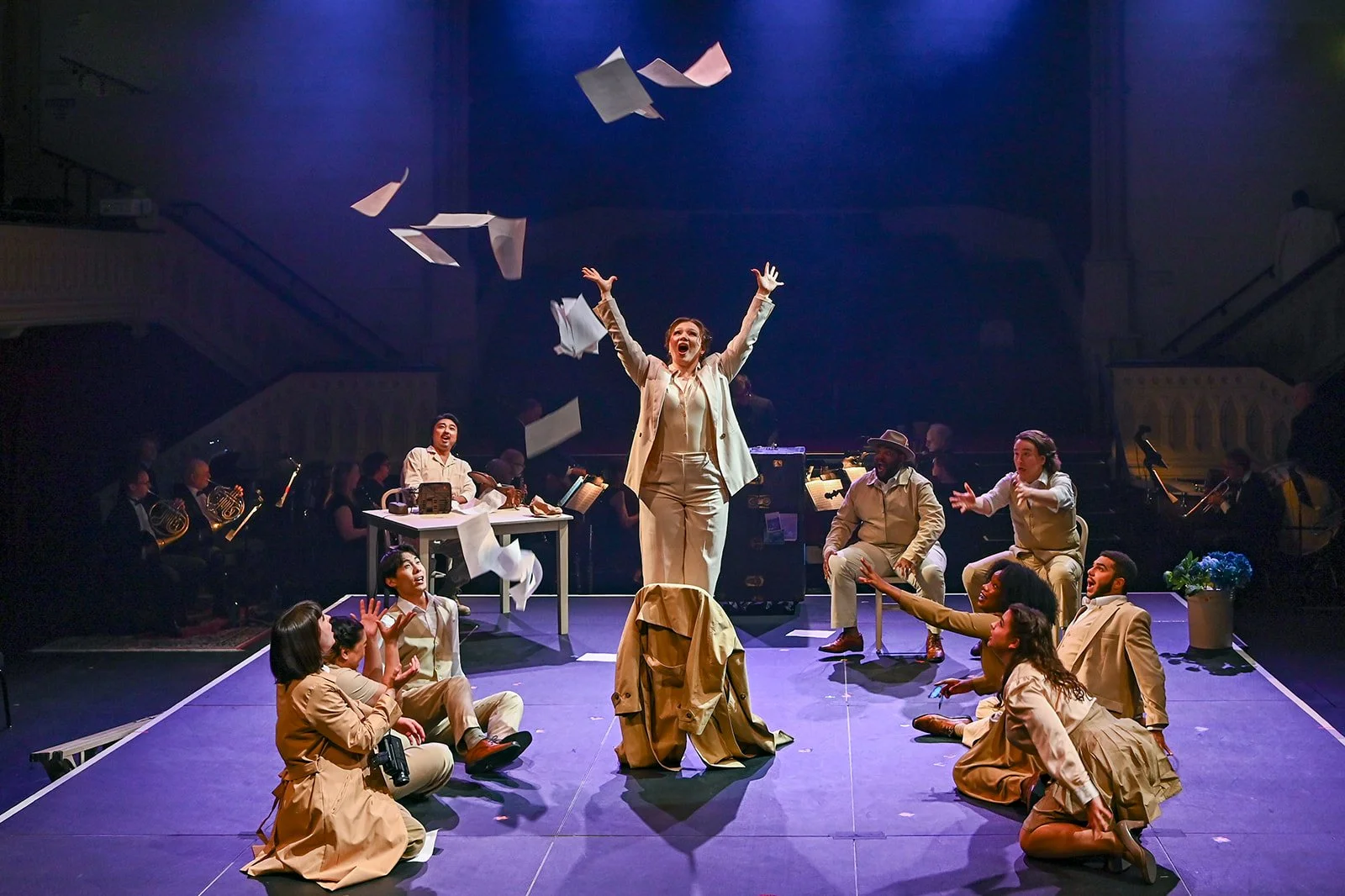 A theatrical performance with actors on stage, table, and musical instruments in the background. The central actress is standing with arms raised, surrounded by seated actors reaching towards her, with papers flying in the air. The scene is lively an