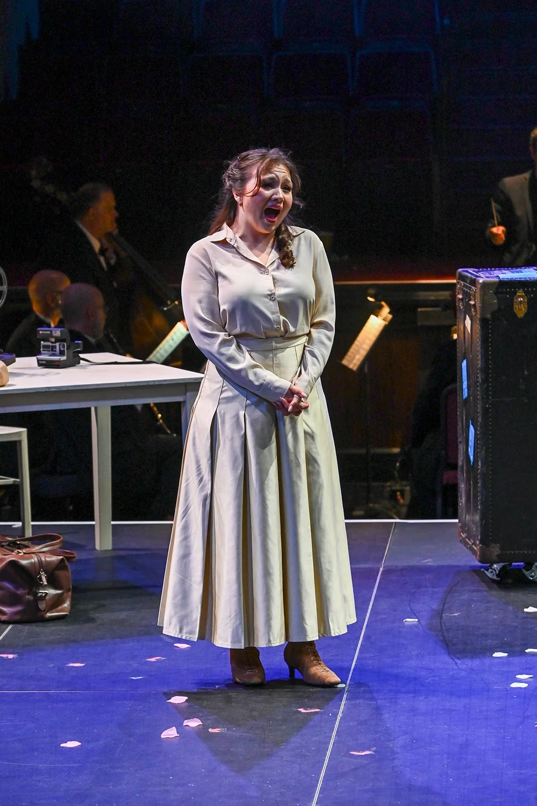 A woman in vintage style clothing with a beige skirt and blouse singing or speaking on stage, with band members and music stands behind her, and scattered rose petals on the floor.