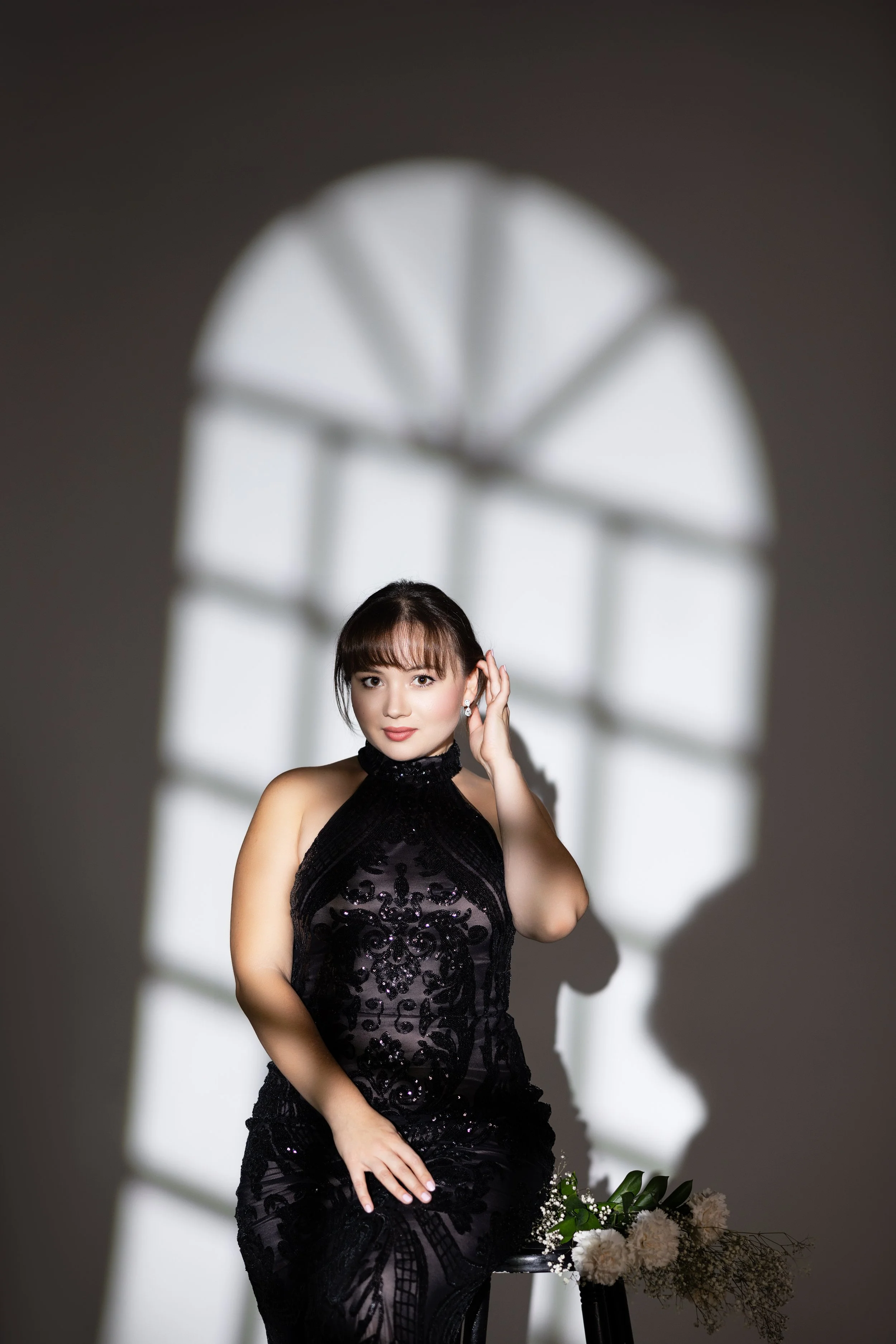 A woman wearing a black, intricately designed dress, standing next to a black stool with a bouquet of white flowers and greenery, in front of a gray wall with a shadow of a window.